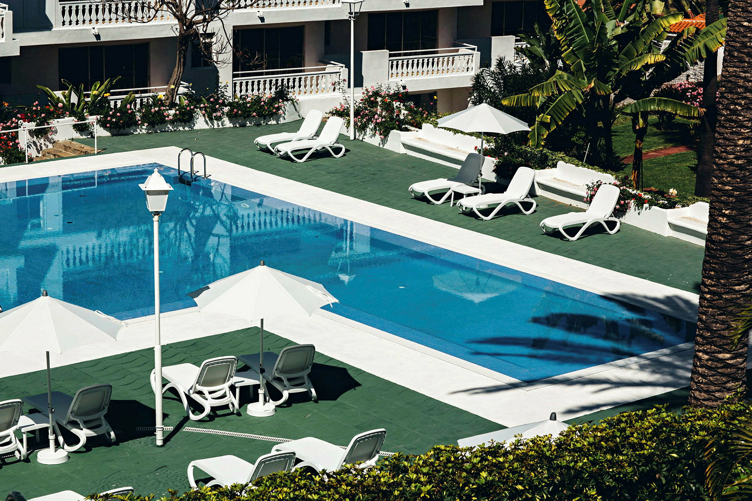 En udendørs pool med liggestole og parasoller omkring, omgivet af grønt og en bygning. (AI-genereret tekst)