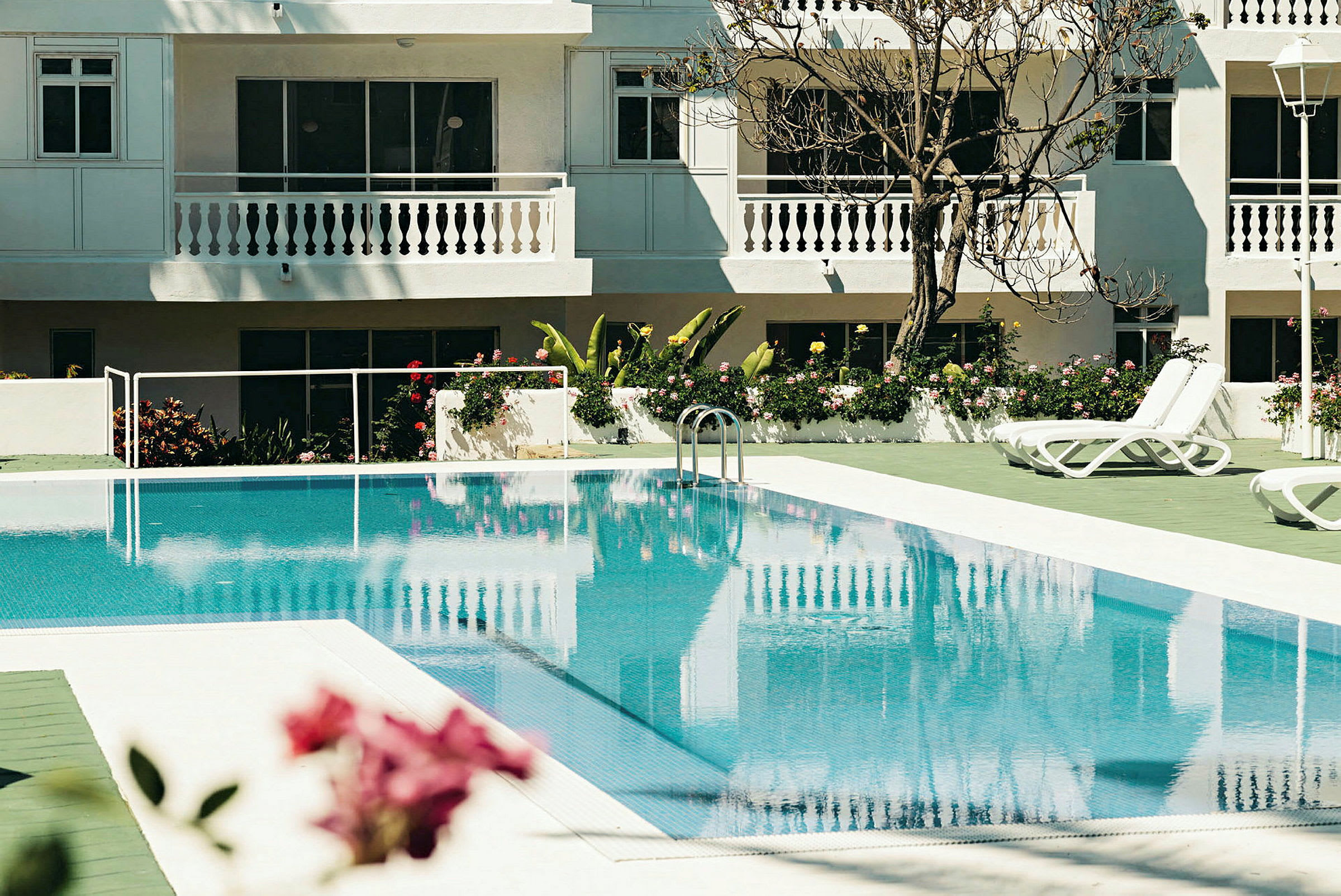 Udendørs swimmingpool med liggestole ved et hvidmalet hotel. (AI-genereret tekst)
