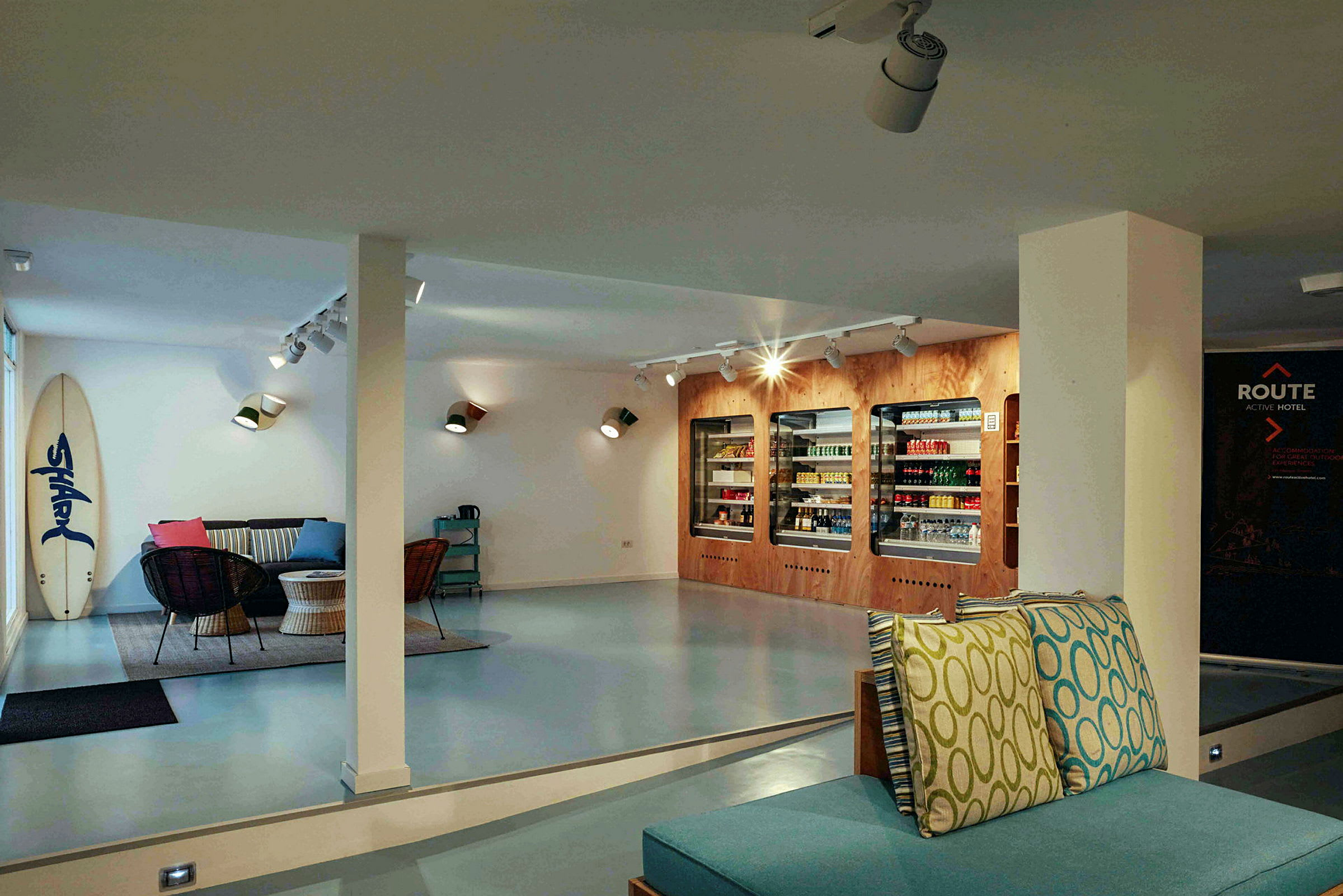 Interiør af en moderne lounge med sofaer, borde og køleskabe fyldt med varer samt et surfbræt, der står op ad væggen. (AI-genereret tekst)