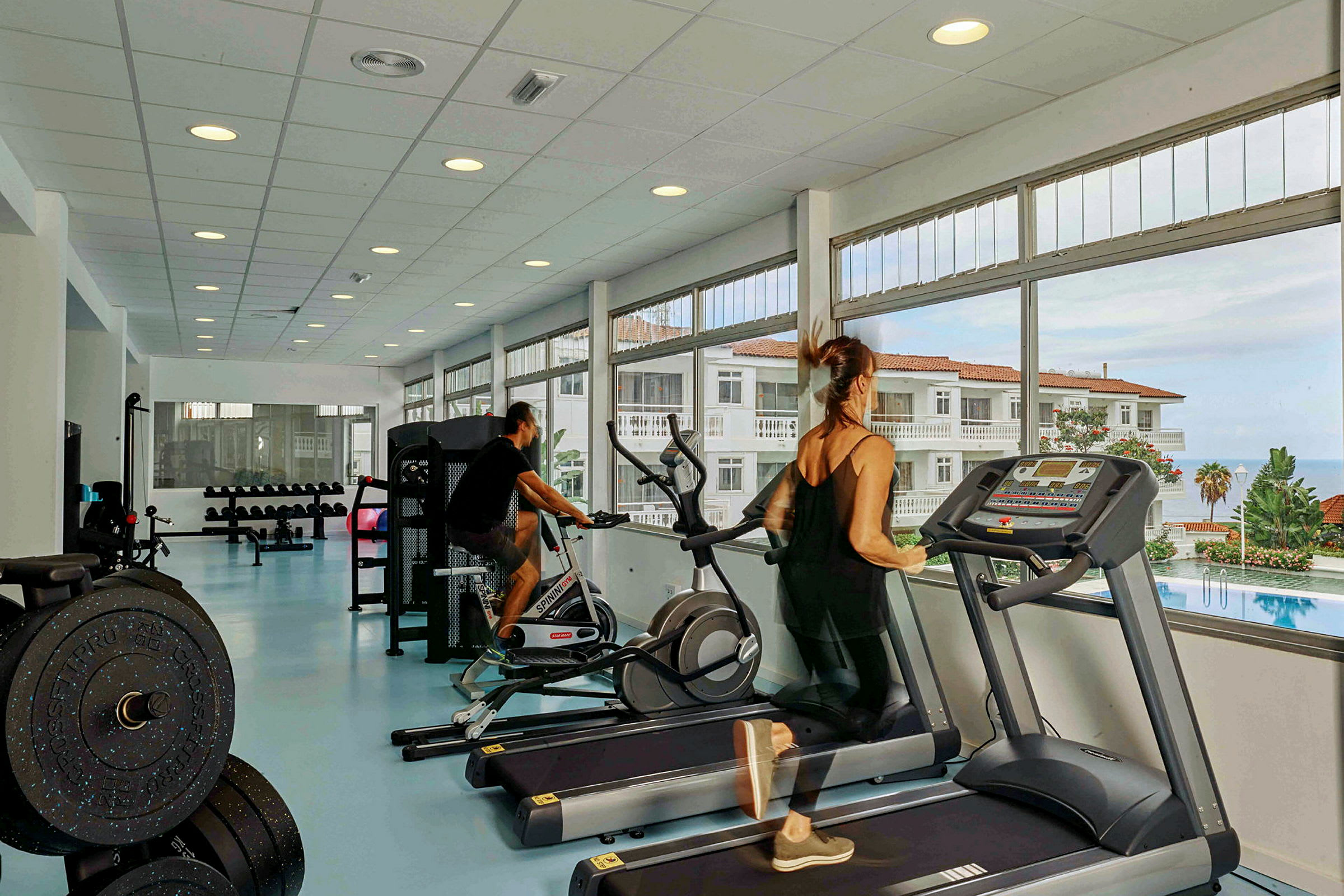 To personer træner i et fitnesscenter med udsigt til en pool og bygninger. (AI-genereret tekst)