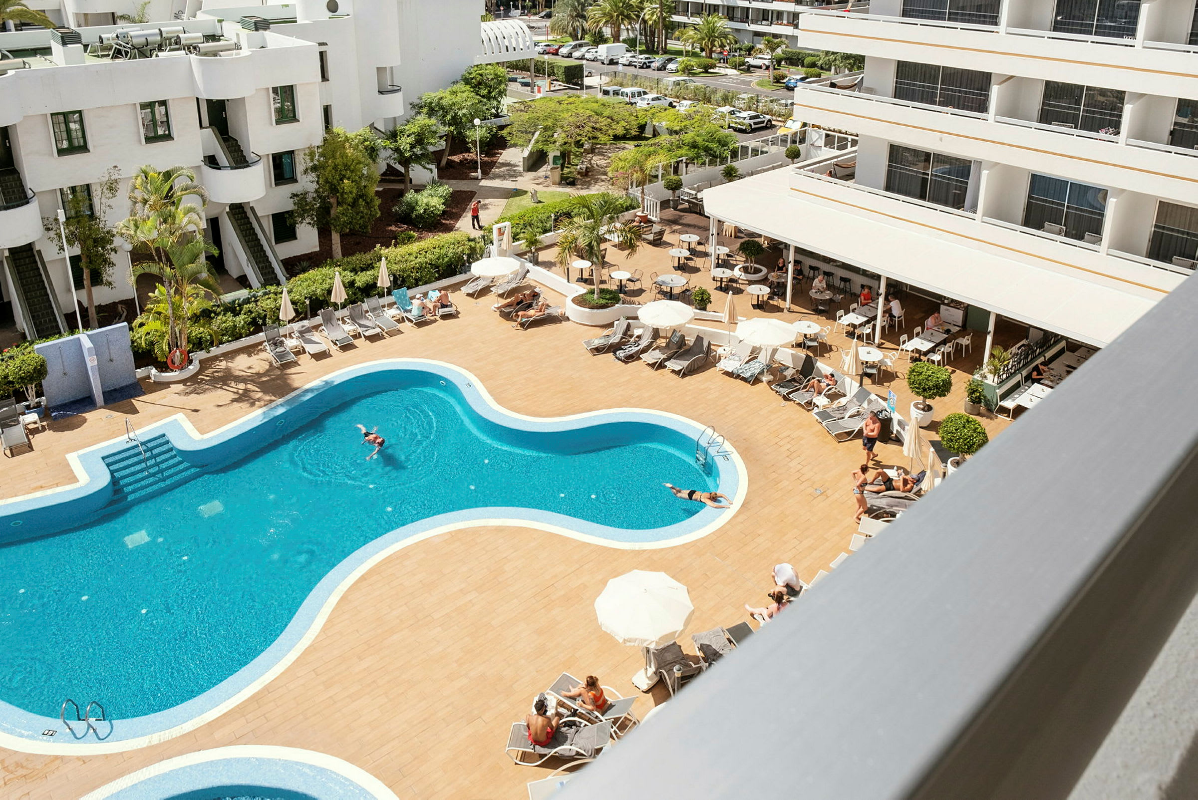 Billede af en hotelpool med liggestole og parasoller, omgivet af hotelbygninger. (AI-genereret tekst)