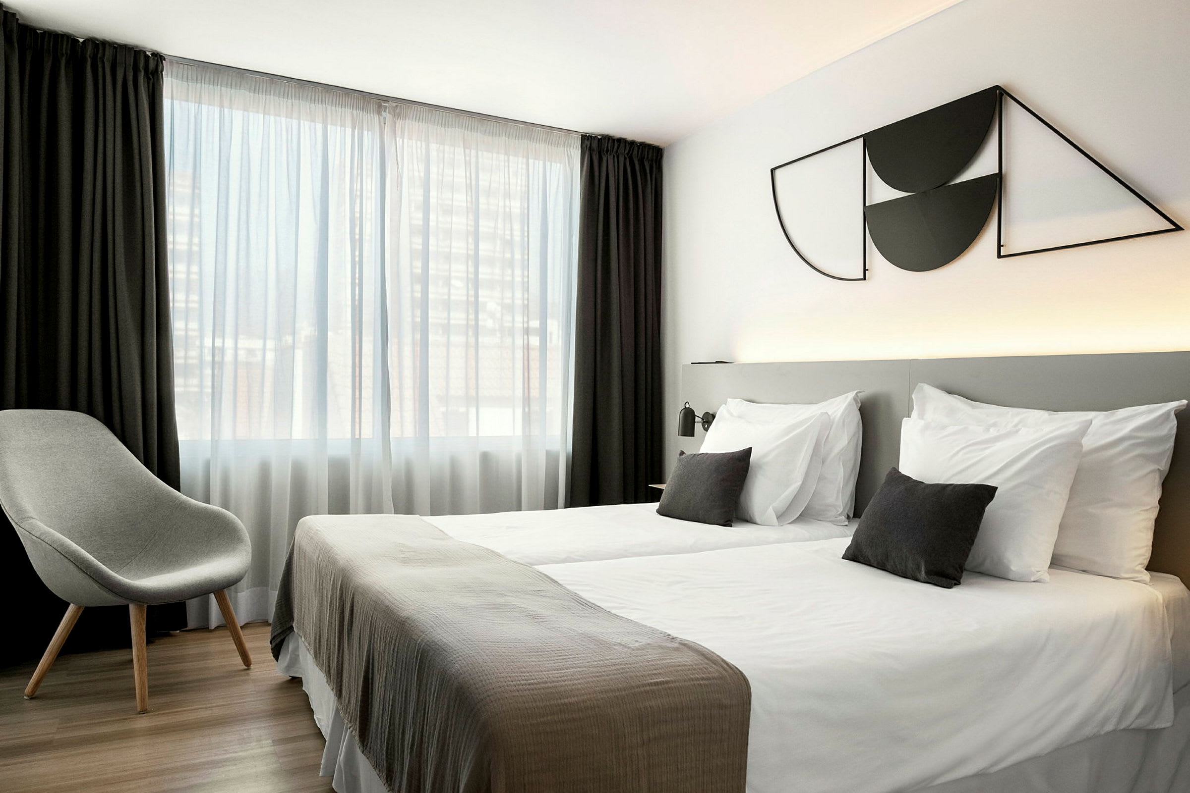 Et hotelværelse med to enkeltsenge, en grå stol og et stort vindue med hvide og mørke gardiner. (AI-genereret tekst)