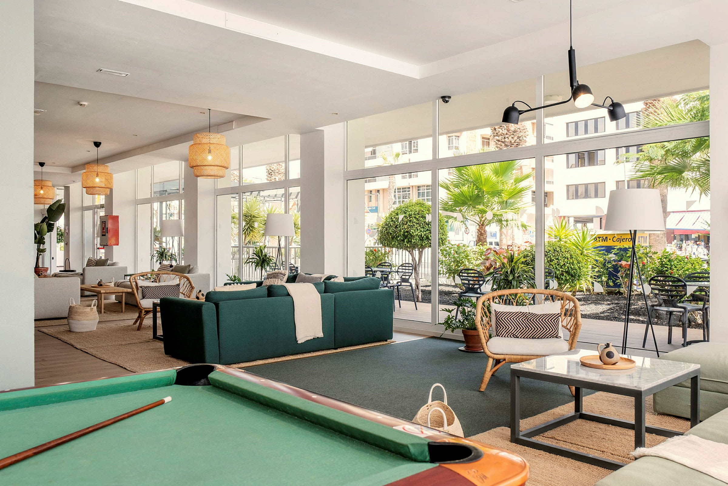 En moderne stue med grønne sofaer, rottingmøbler og et billardbord i forgrunden. Store vinduer vender ud mod en terrasse med planter og træer. (AI-genereret tekst)