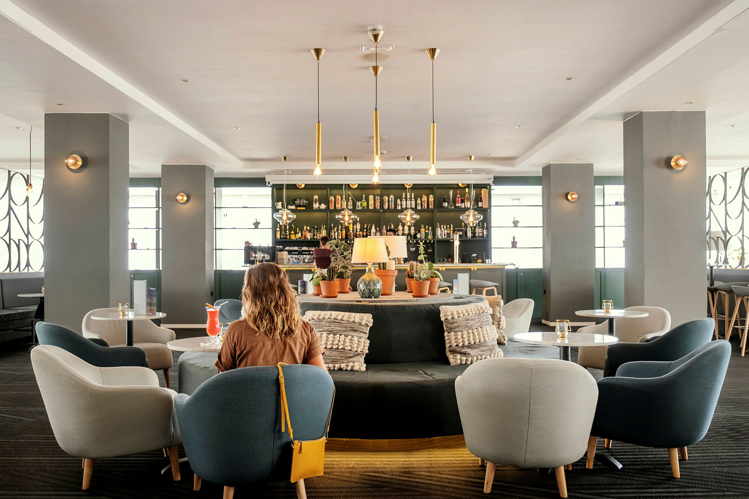 Interiør af et moderne hotelbarområde med siddepladser og dekorationer. En kvinde med ryggen til kameraet i forgrunden. (AI-genereret tekst)