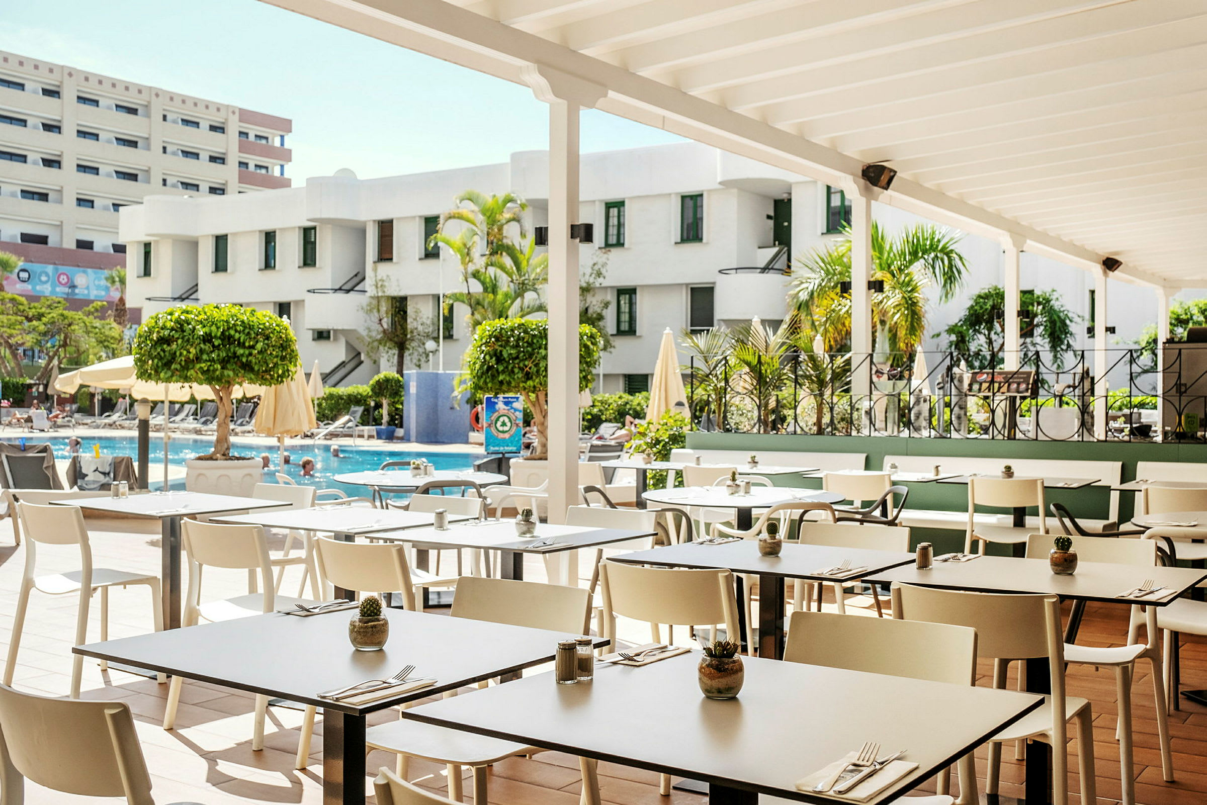 En restaurantterrasse med udsigt over poolområdet omgivet af bygninger og grønt. (AI-genereret tekst)
