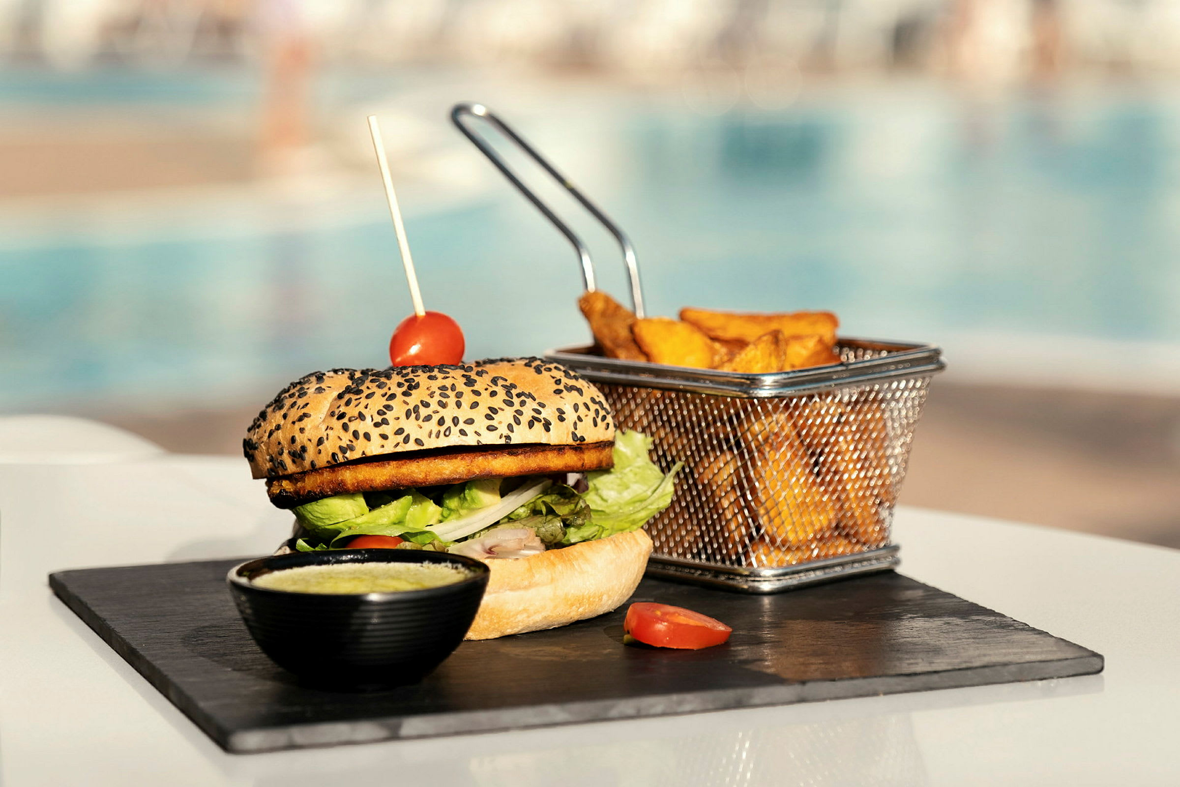 En burger med tomatdekoration, ved siden af pommes frites i en metalkurv, serveret på en stenplade. (AI-genereret tekst)