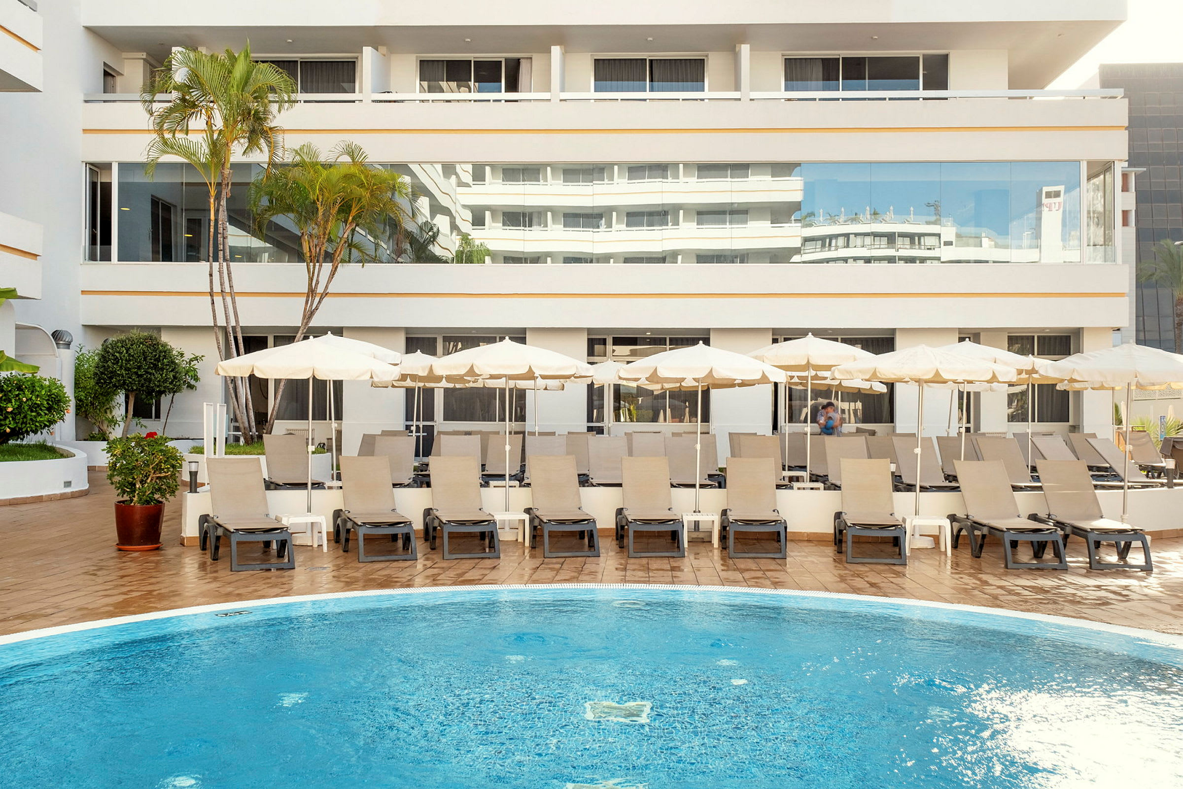 Billede af en hotelpool med liggestole og parasoller foran en lys bygning. (AI-genereret tekst)
