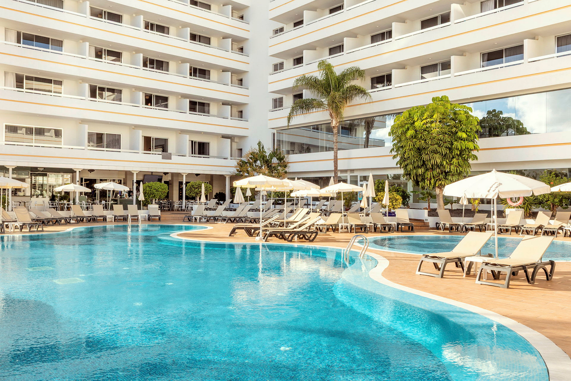 Billede af en hotelpool med liggestole og parasoller ved siden af og et fler-etagers hotel i baggrunden. (AI-genereret tekst)