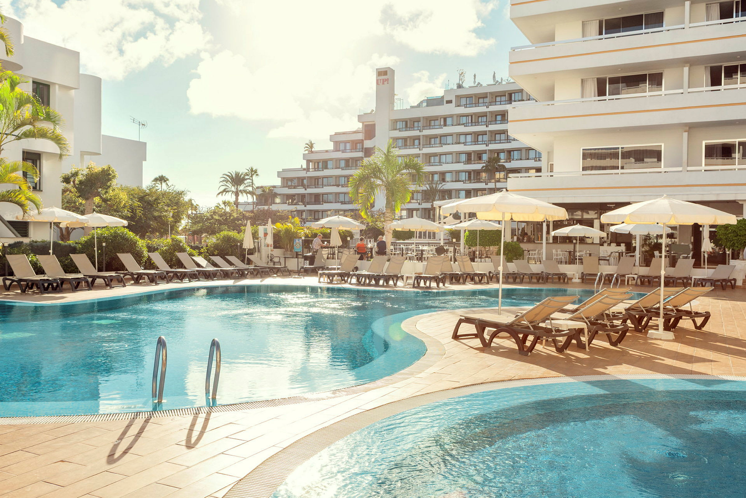 Hotelpool med liggestole og parasoller, omgivet af bygninger. (AI-genereret tekst)