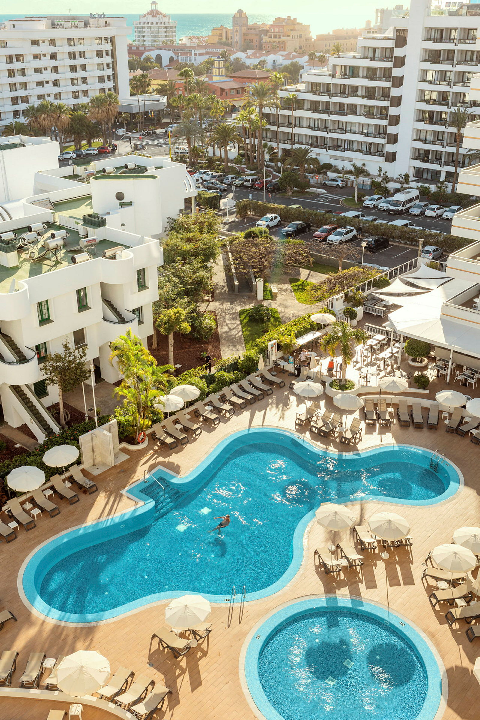 Billede af et hotelområde med pools og liggestole, omgivet af grønne planter og bygninger. (AI-genereret tekst)