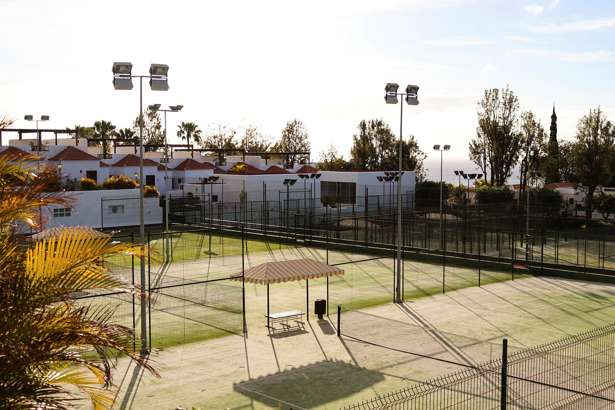 Billede af en padeltennisbane omkranset af hegn, med udsigt over en bygning med hvide vægge og røde tage. (AI-genereret tekst)