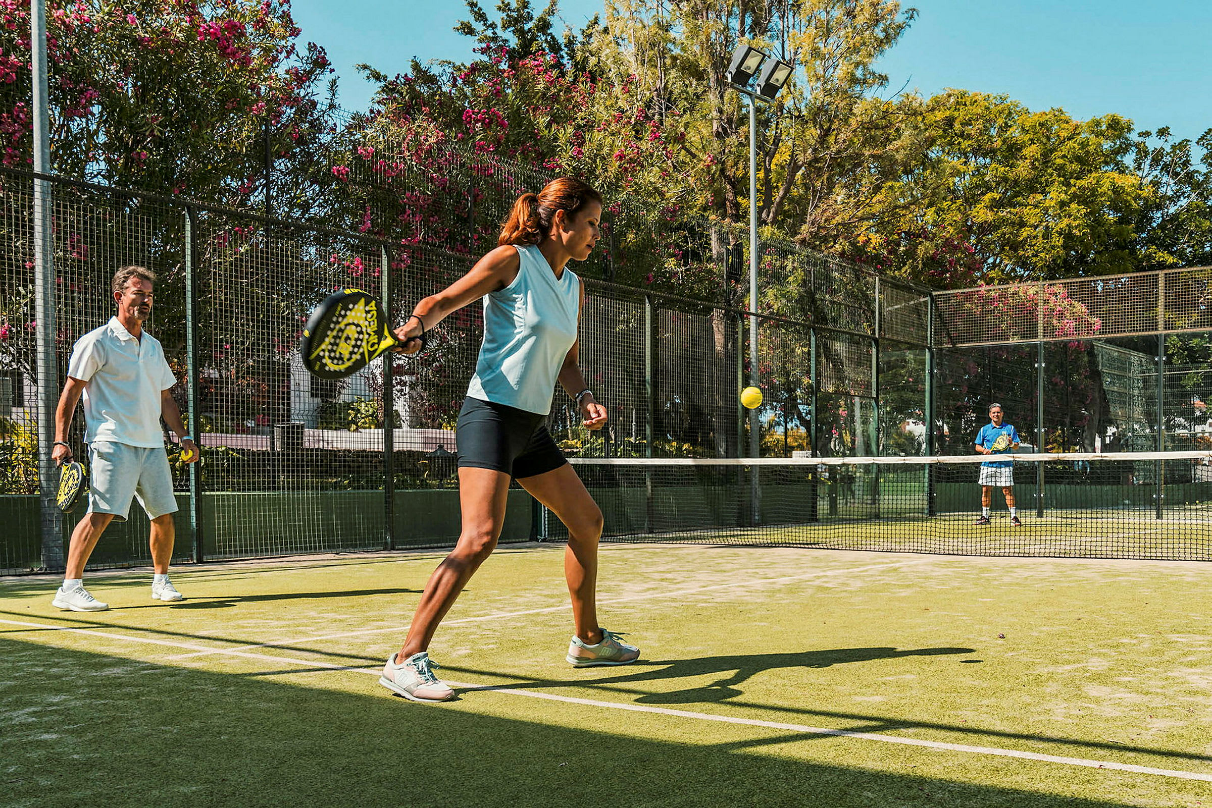 Personer spiller padel på en udendørs bane omgivet af grønne omgivelser. (AI-genereret tekst)