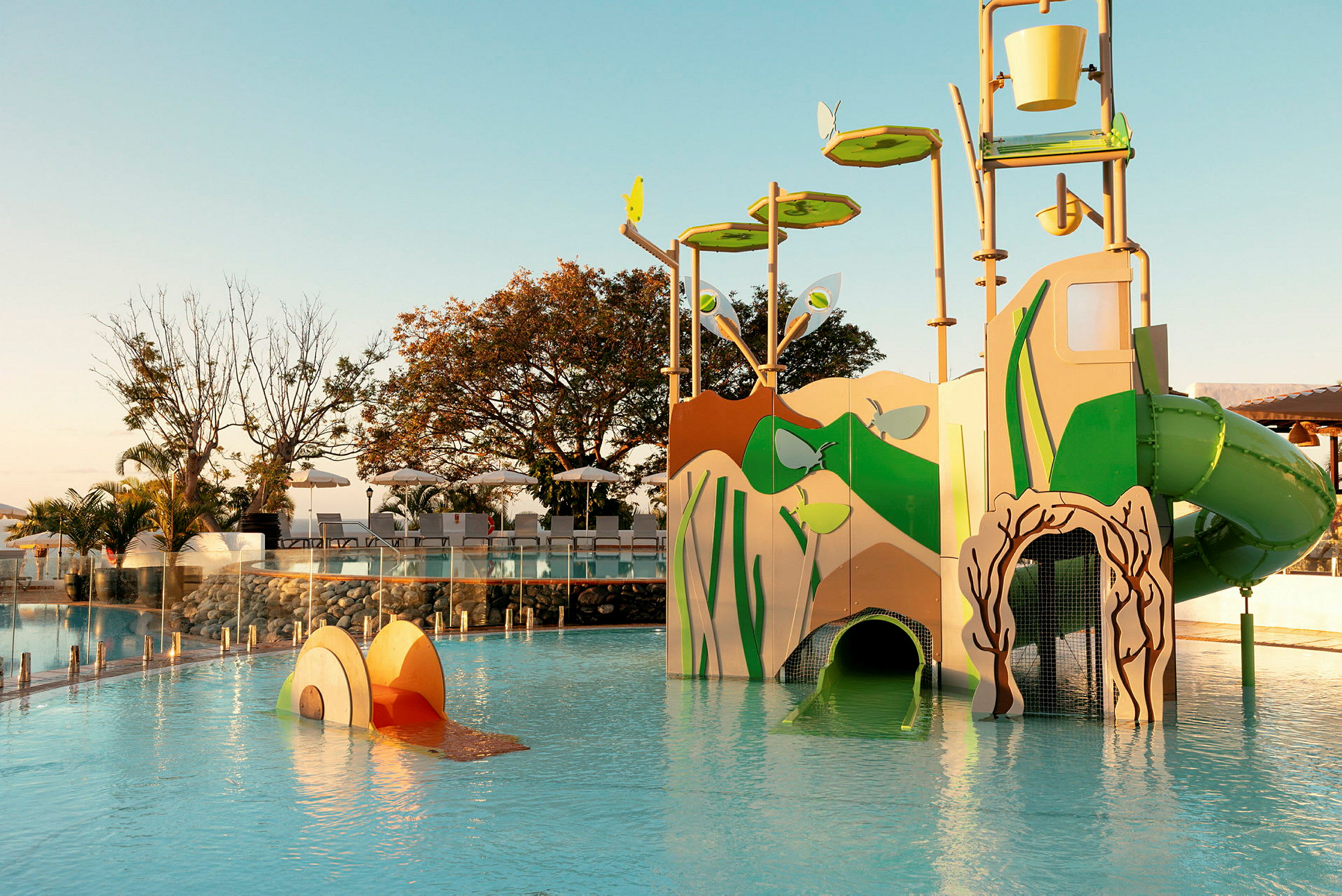 En vandlegeplads med en struktur, der inkluderer rutsjebaner og interaktive elementer, omgivet af swimmingpool og liggestole. (AI-genereret tekst)