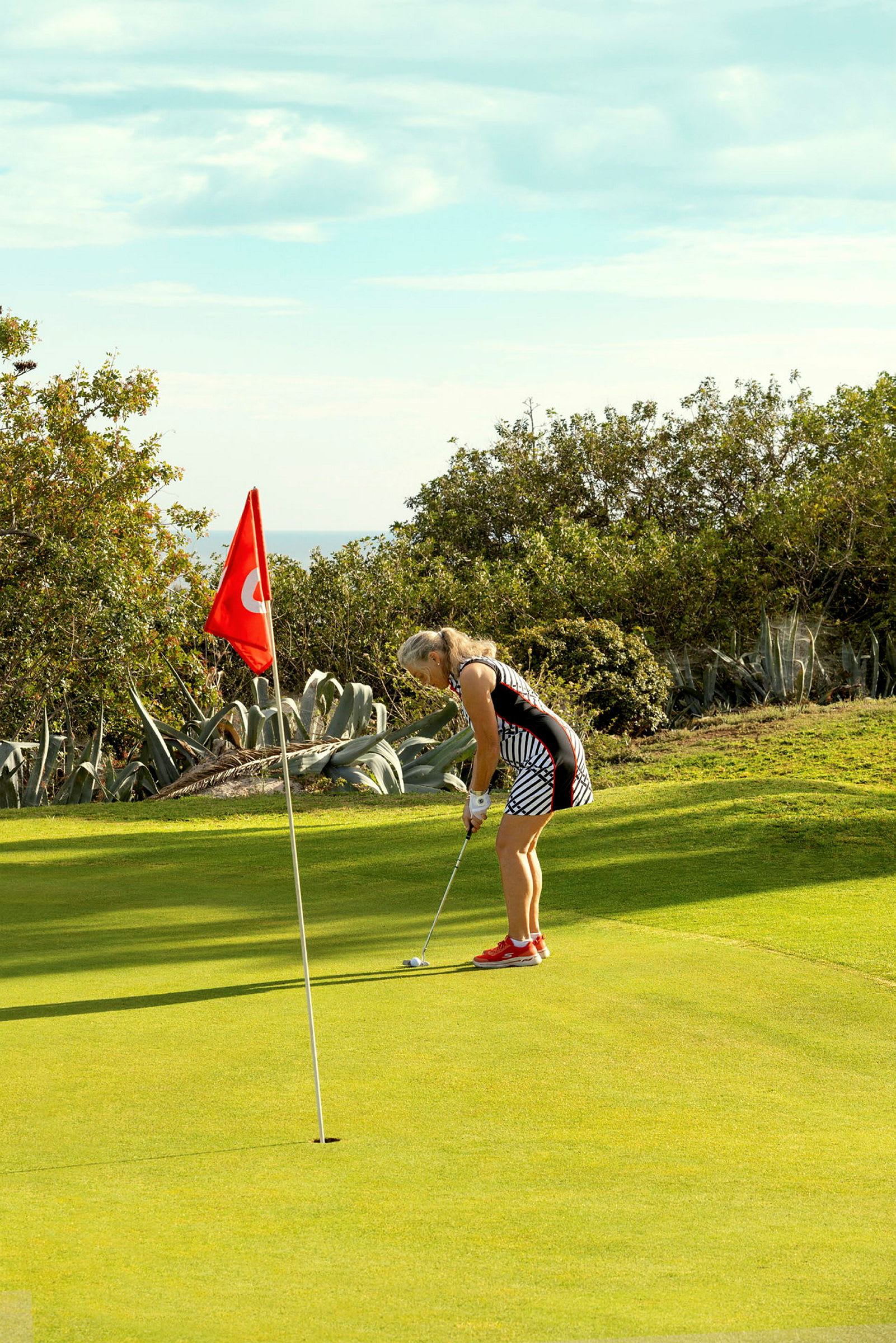 En person spiller golf og sigter mod hullet nær en rød flag, med grøn natur i baggrunden. (AI-genereret tekst)