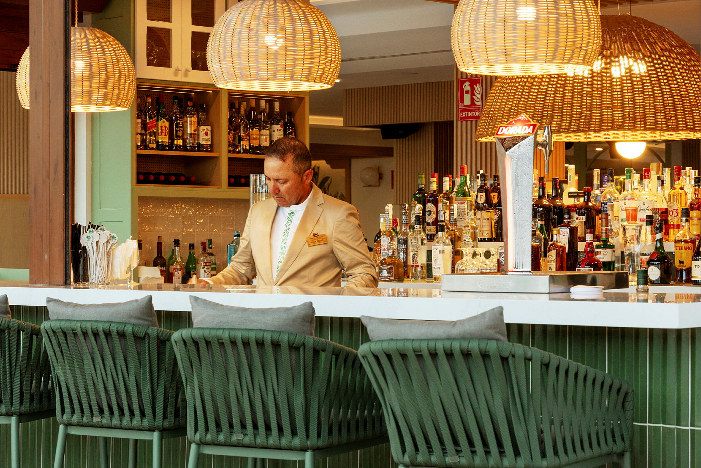 Interiør af en elegant bar med flettede lamper og stole samt et udvalg af flasker og drikkevarer på hylderne. (AI-genereret tekst)