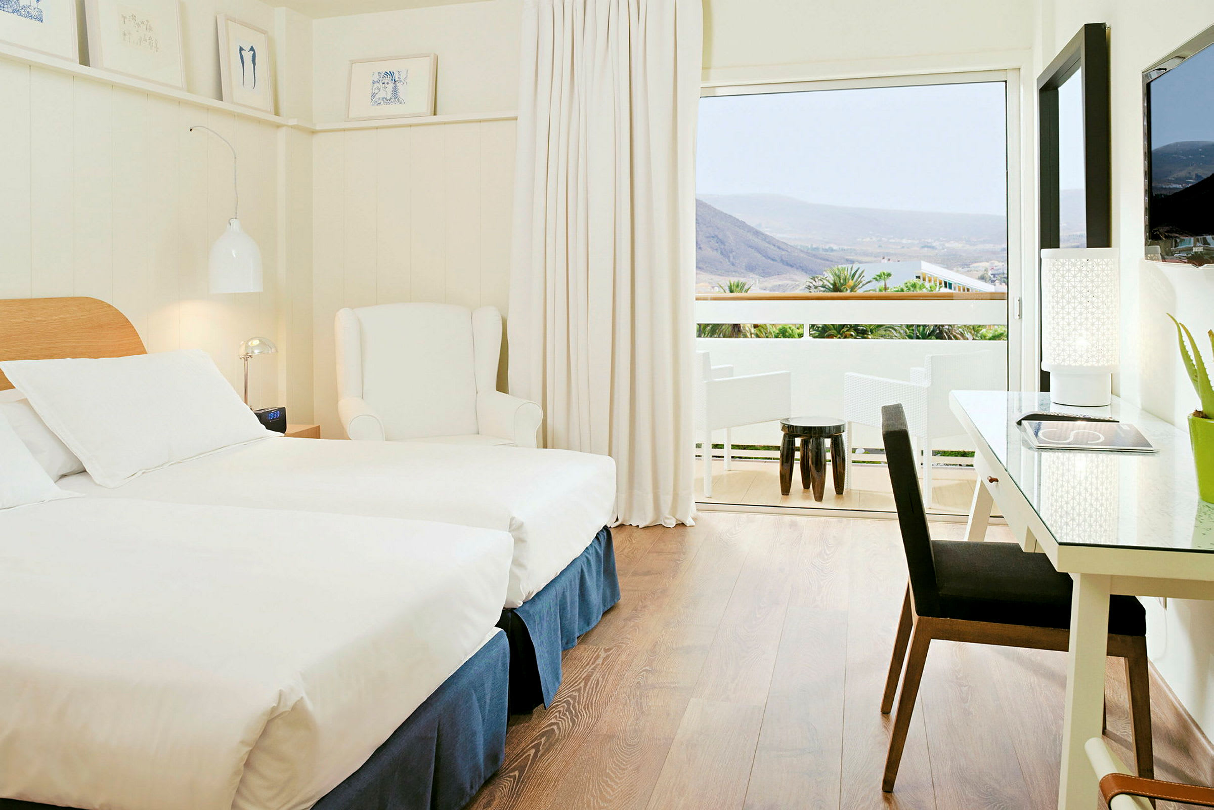 Et lyst og moderne hotelværelse med to enkeltsenge, en hvid lænestol og et skrivebord med lampe. Et stort vindue fører ud til en balkon med udsigt over bjerge og grønt. (AI-genereret tekst)