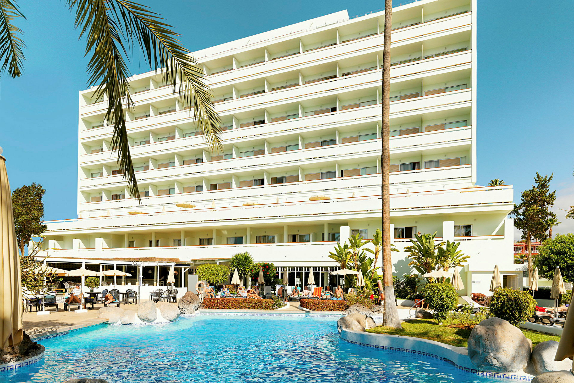 Et hotel med flere etager og altaner ved en udendørs pool omgivet af liggestole og parasoller. (AI-genereret tekst)