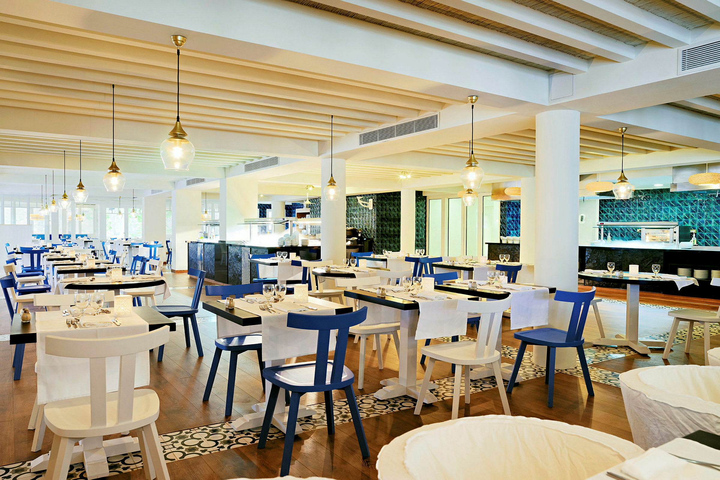 En lys og moderne restaurant med hvide og blå stole og borde dækket til middag. Loftet har synlige træbjælker og hængende lamper. Mønstret gulv og køkkenområde i baggrunden. (AI-genereret tekst)