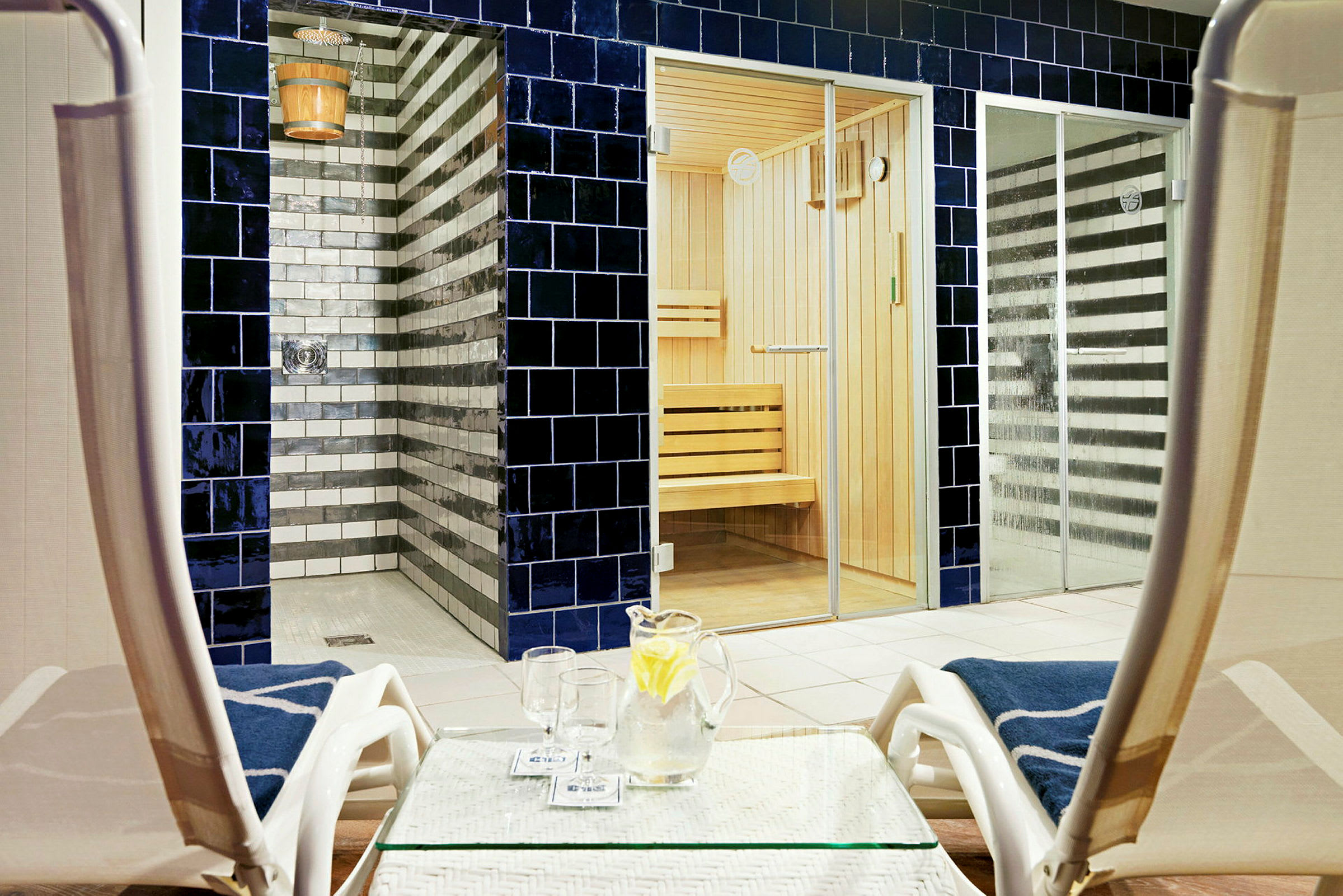 Billede af et spaområde med sauna og brusere i baggrunden, samt solsenge og et bord med en kande vand og glas i forgrunden. (AI-genereret tekst)