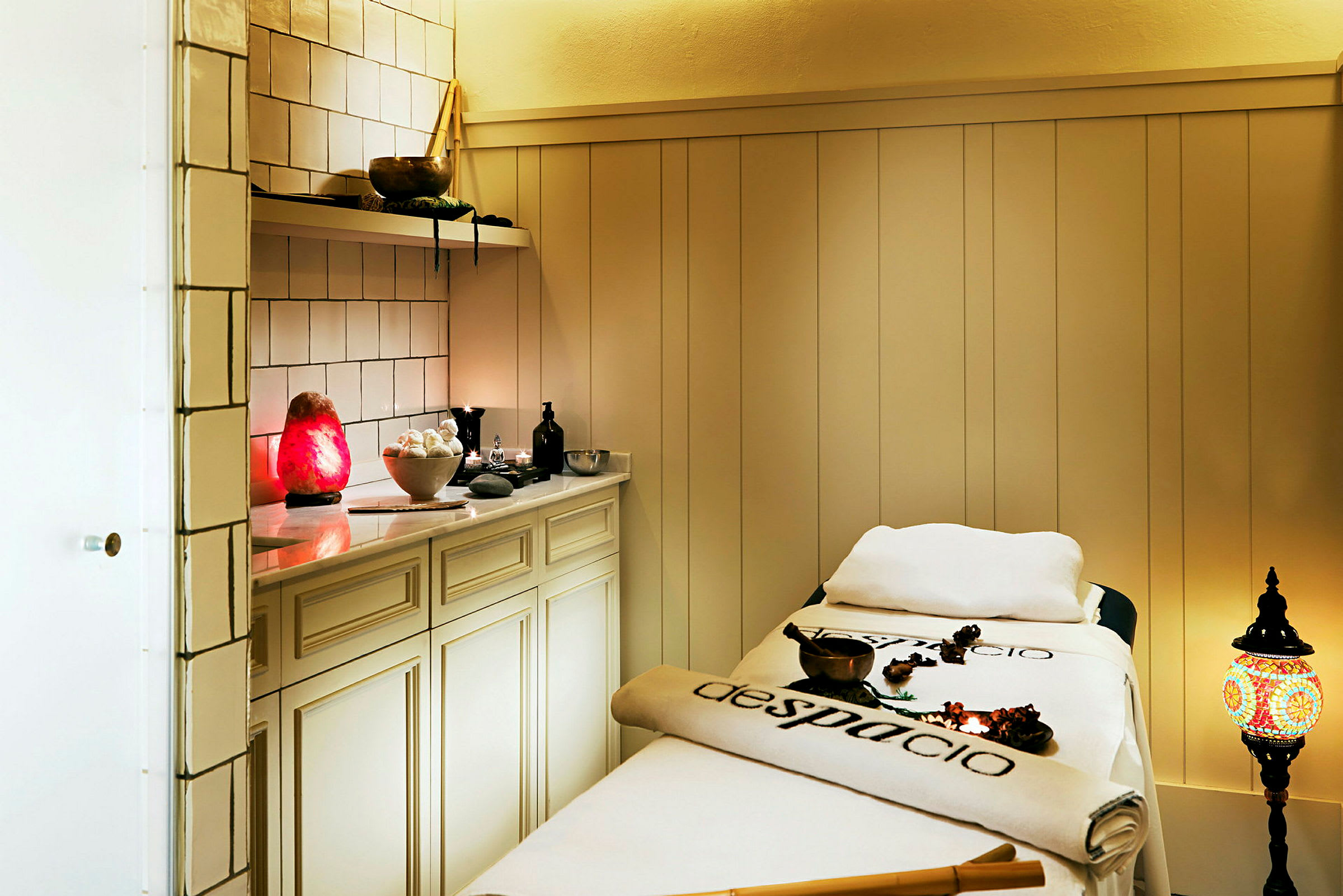 Billede af et spa-rum med behandlingsseng, skab og dekorative genstande, herunder en rød lampe og en farverig lanterne. (AI-genereret tekst)