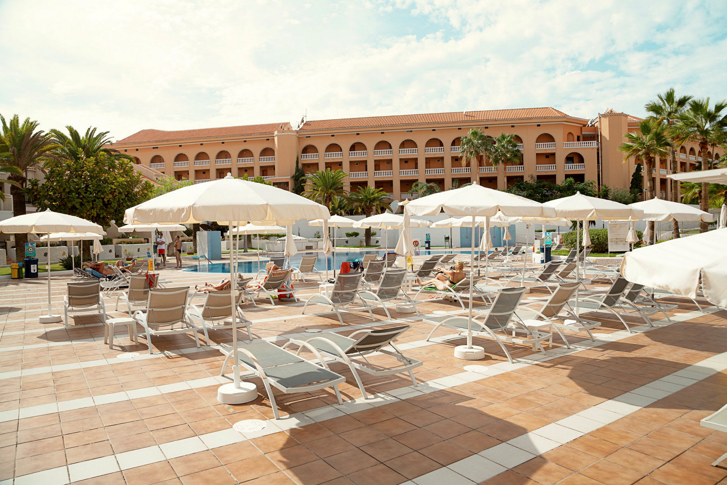 Liggestole under parasoller på en terrasse tæt på en pool ved en hotelbygning. (AI-genereret tekst)
