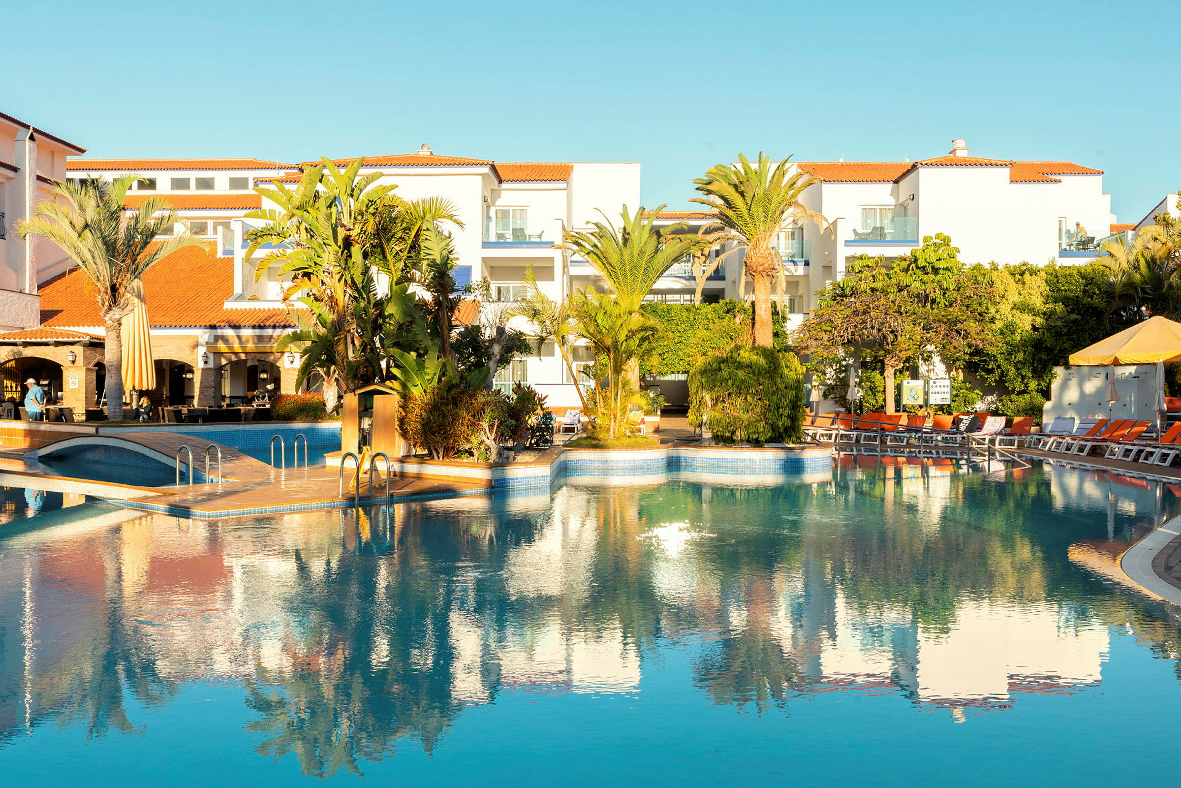 En ferieudvej med huse i hvide og orange farver ved siden af en udendørs pool omgivet af frodige planter og liggestole. (AI-genereret tekst)