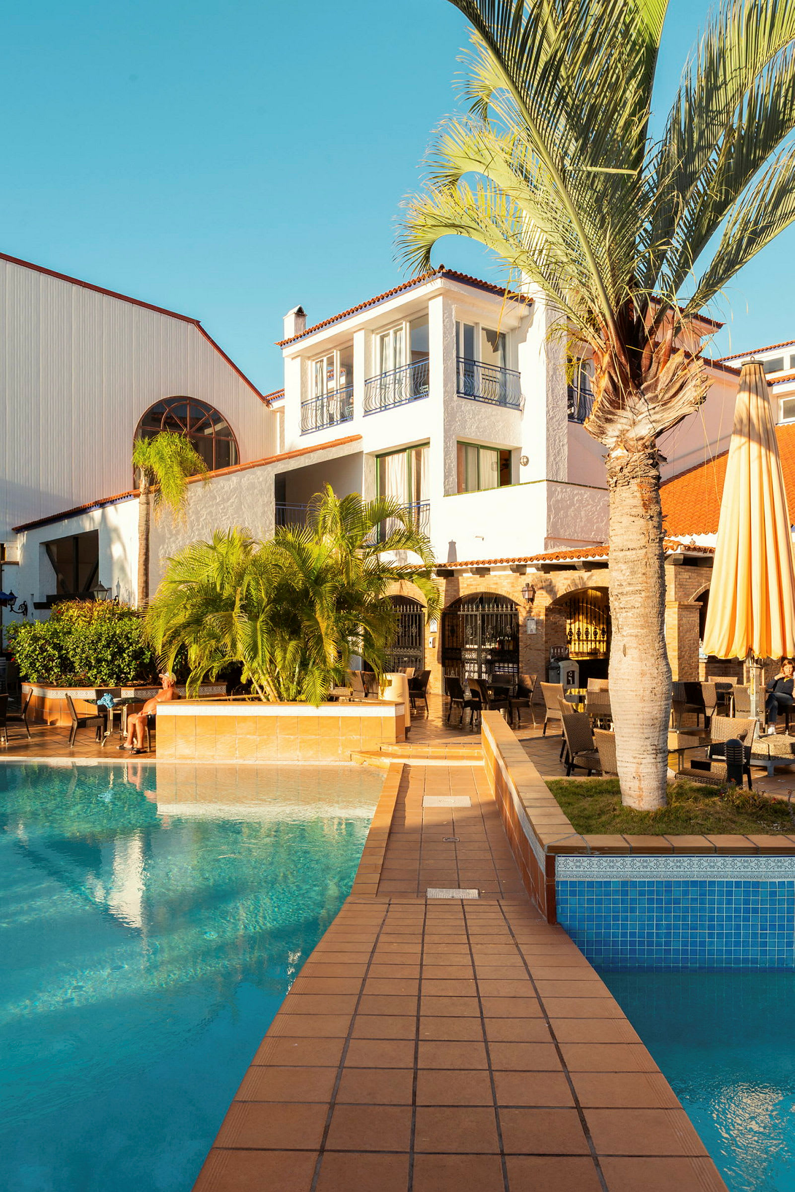 En bygning med hvid facade og altaner tæt på en pool med terrasse. (AI-genereret tekst)