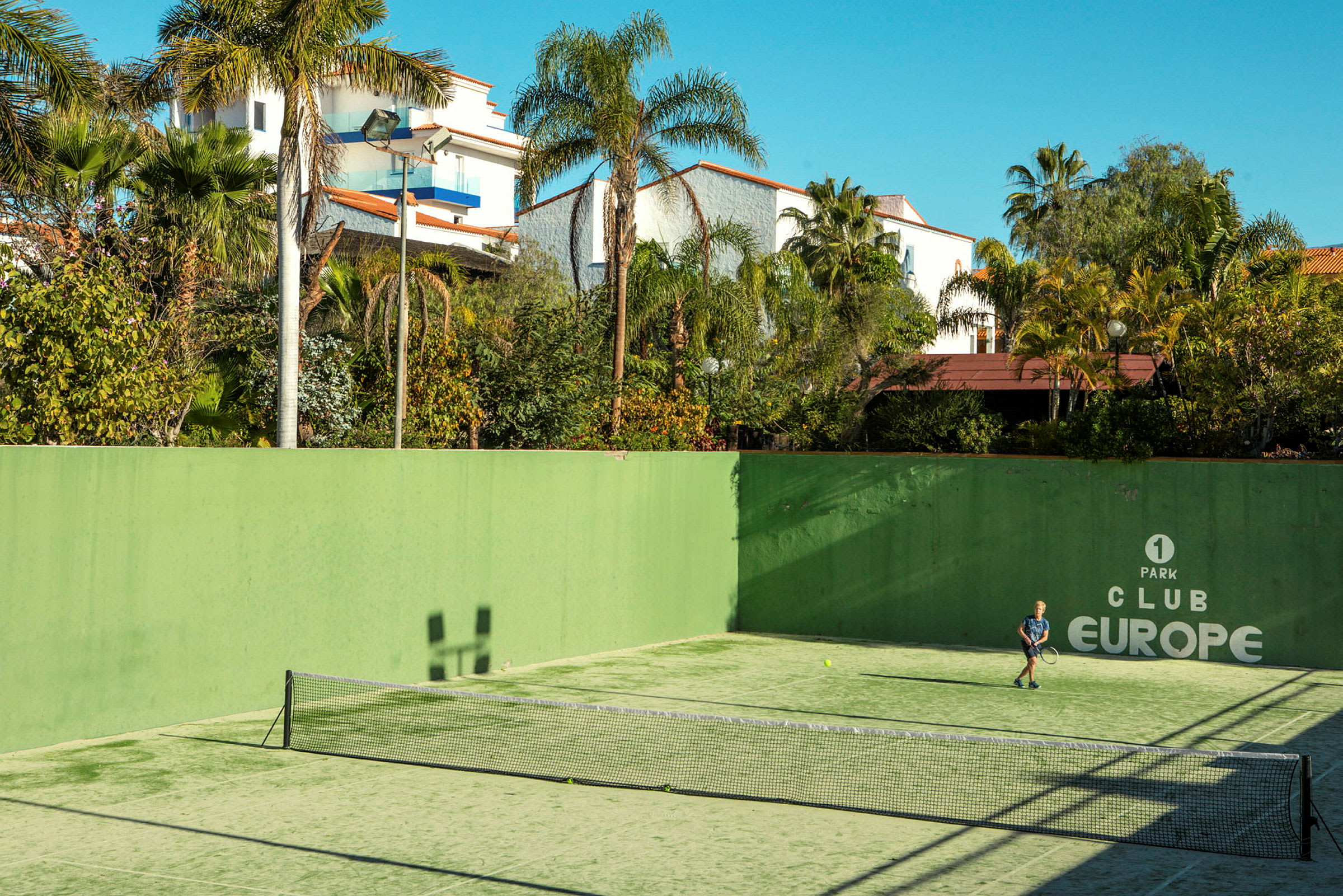 En udendørs tennisbane med en person, der spiller tennis, omgivet af grønne planter og bygninger. (AI-genereret tekst)