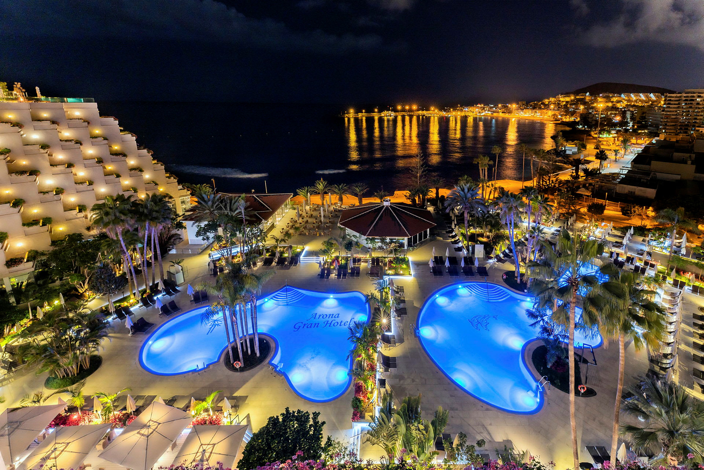 Natudsigt over et hotel med oplyste swimmingpools og palmer ved havets kyst. (AI-genereret tekst)