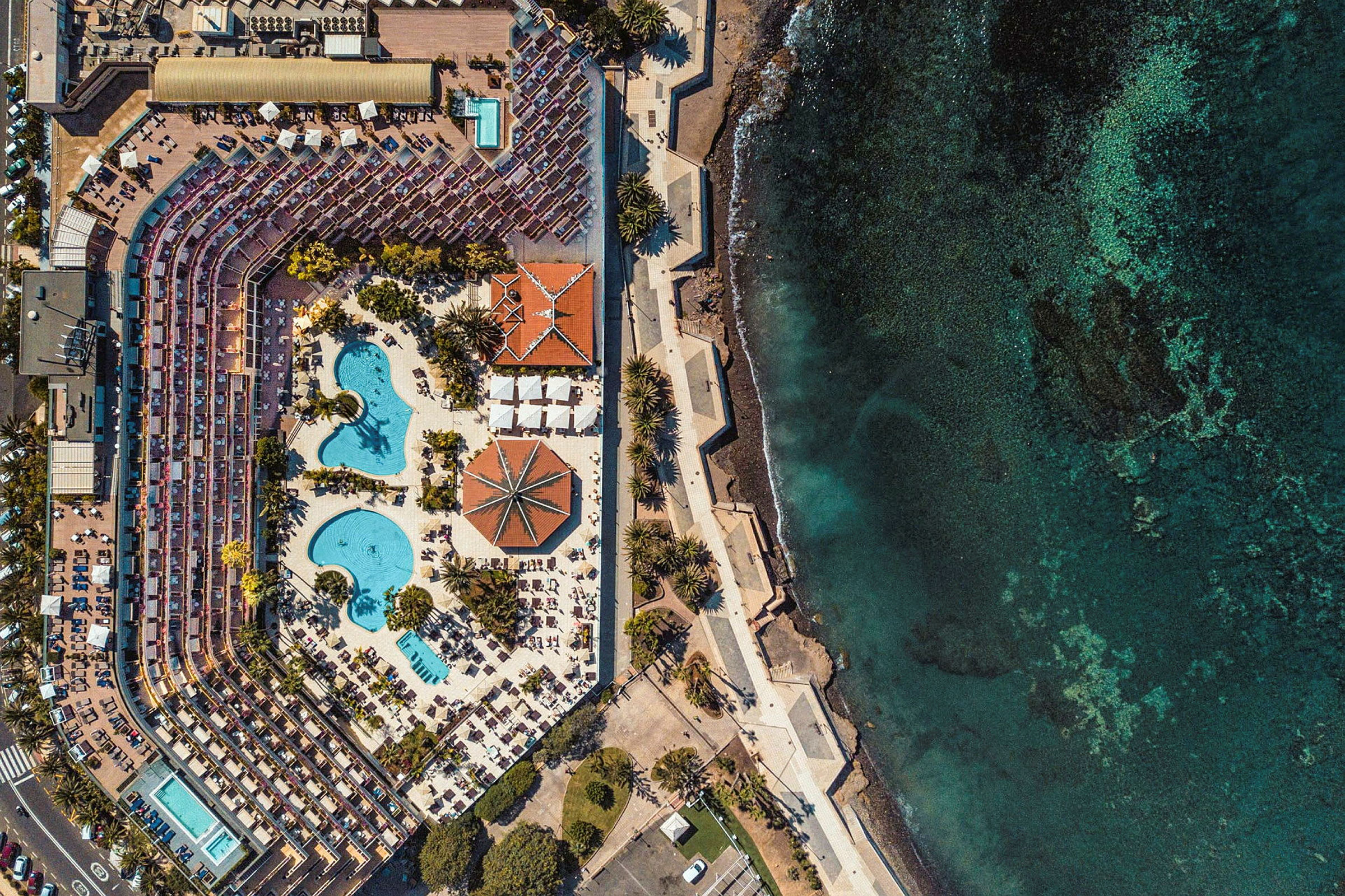 Luftfoto af hotelområde med pools, liggestole og hav ved siden. (AI-genereret tekst)