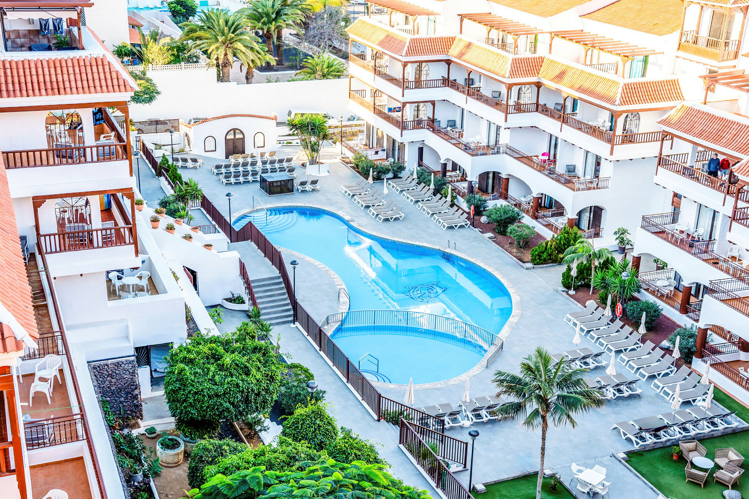 Hotellets poolområde med liggestole og omliggende bygninger. (AI-genereret tekst)