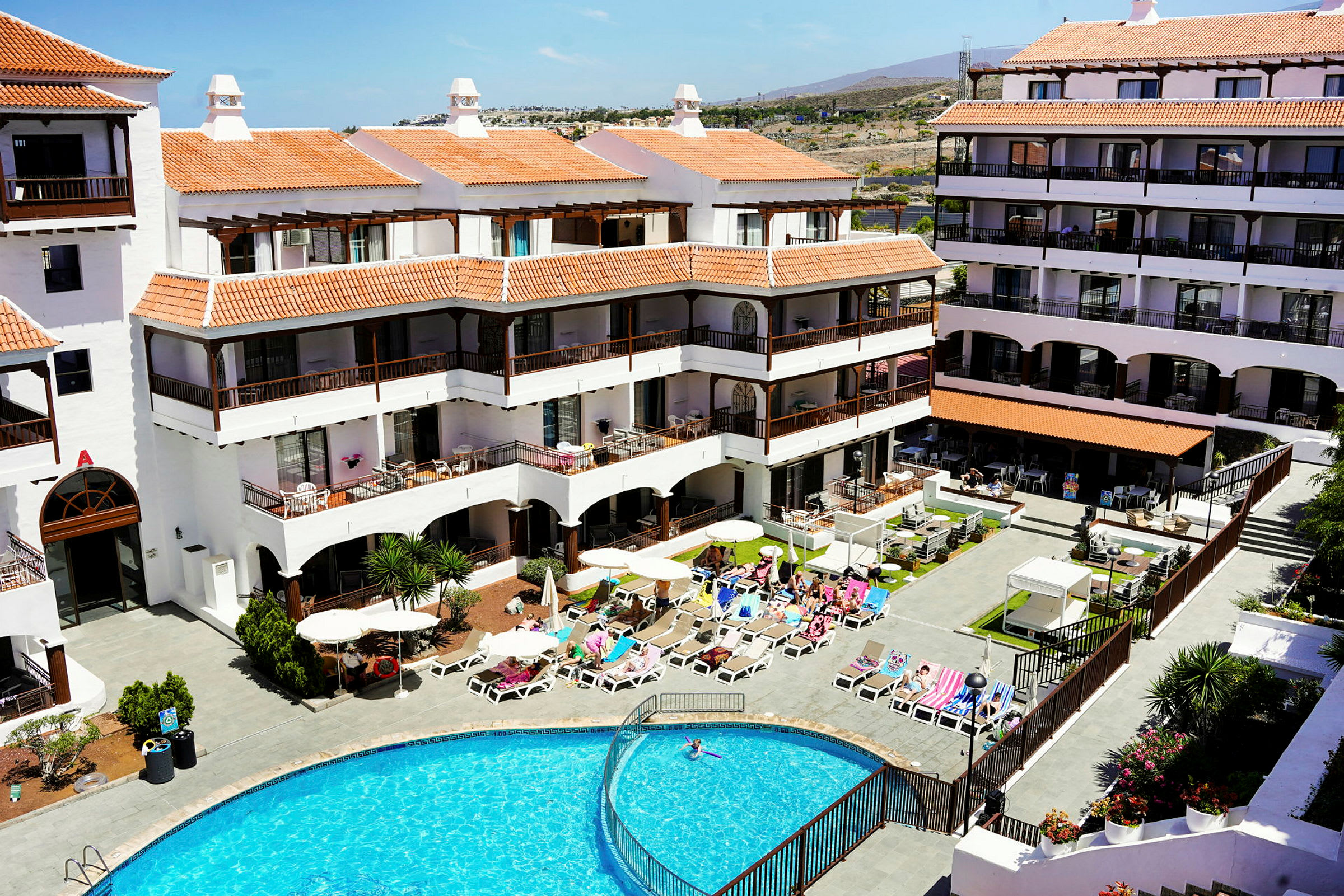 Hotel med hvidmalet facade, terrakottafarvet tag og flere etager med altaner omkring en udendørs pool. (AI-genereret tekst)
