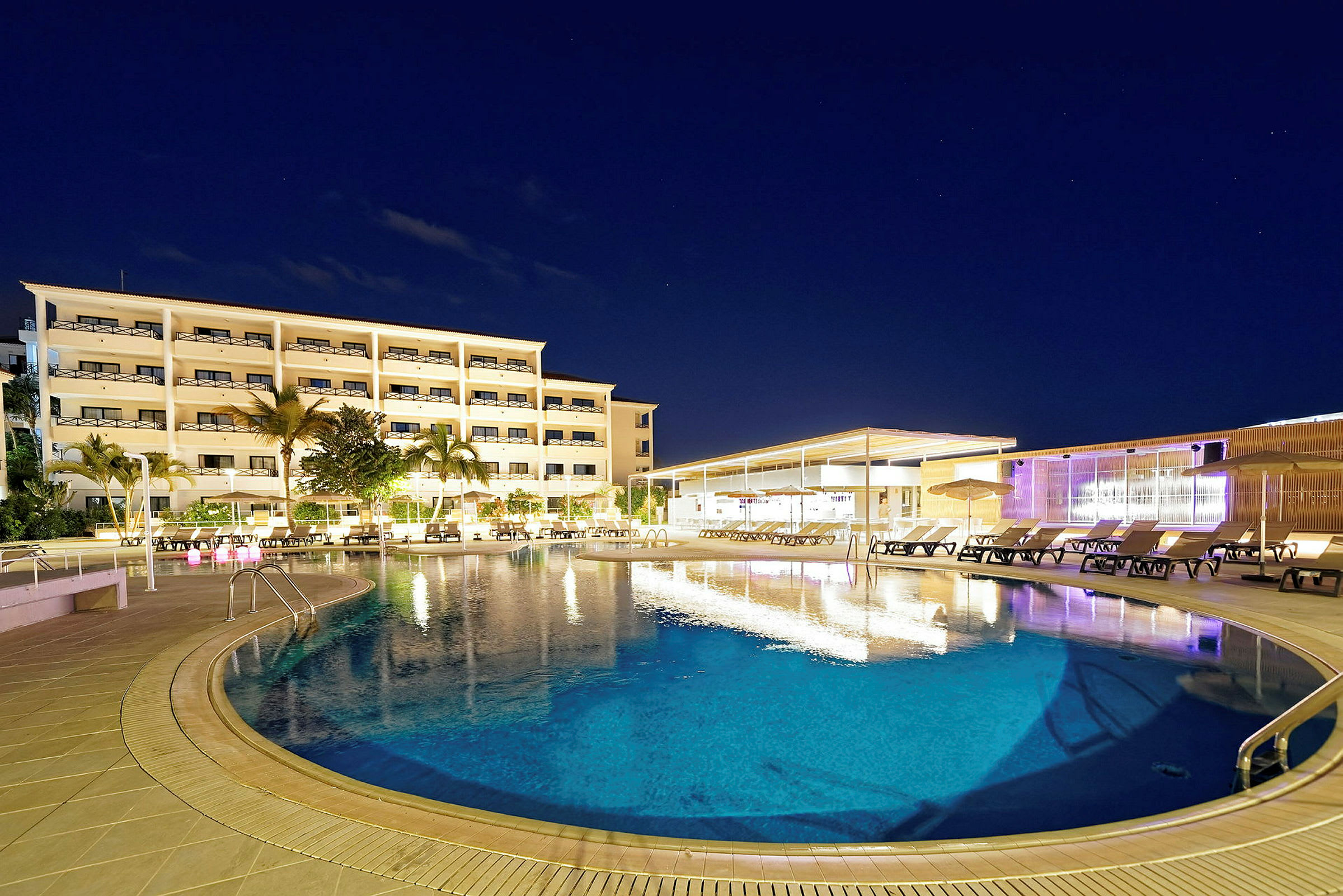 Et hotel med flere etager og en oplyst swimmingpool foran, omgivet af liggestole og palmer, under en nattehimmel. (AI-genereret tekst)