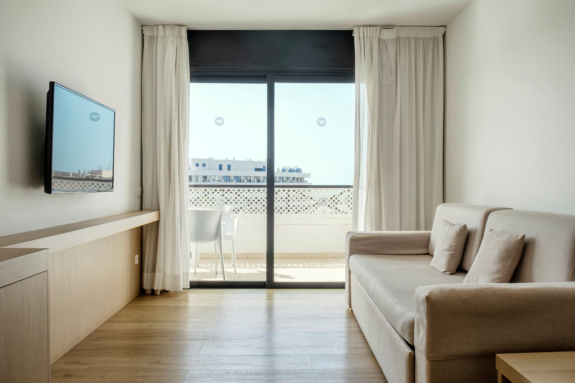 Et lyst og moderne hotelværelse med sofa, vægmonteret TV og stort vindue med udsigt til en altan. (AI-genereret tekst)