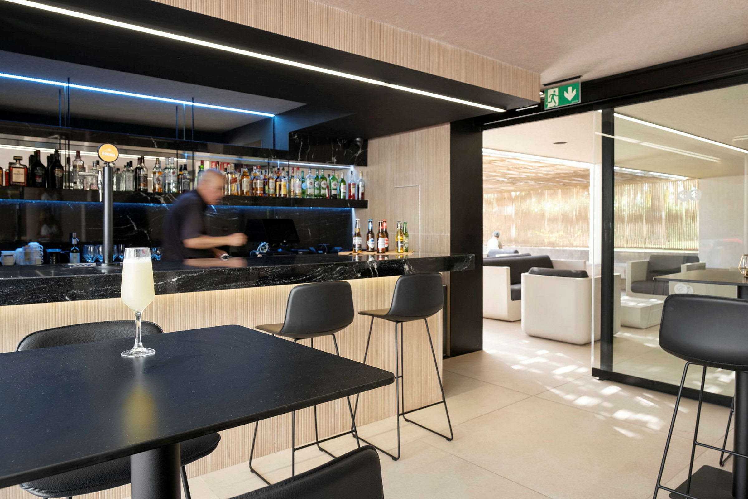Et billede af en bar med en bartender, der laver drikkevarer. Foran ham står stole, og en perlende drink står på et bord. (AI-genereret tekst)