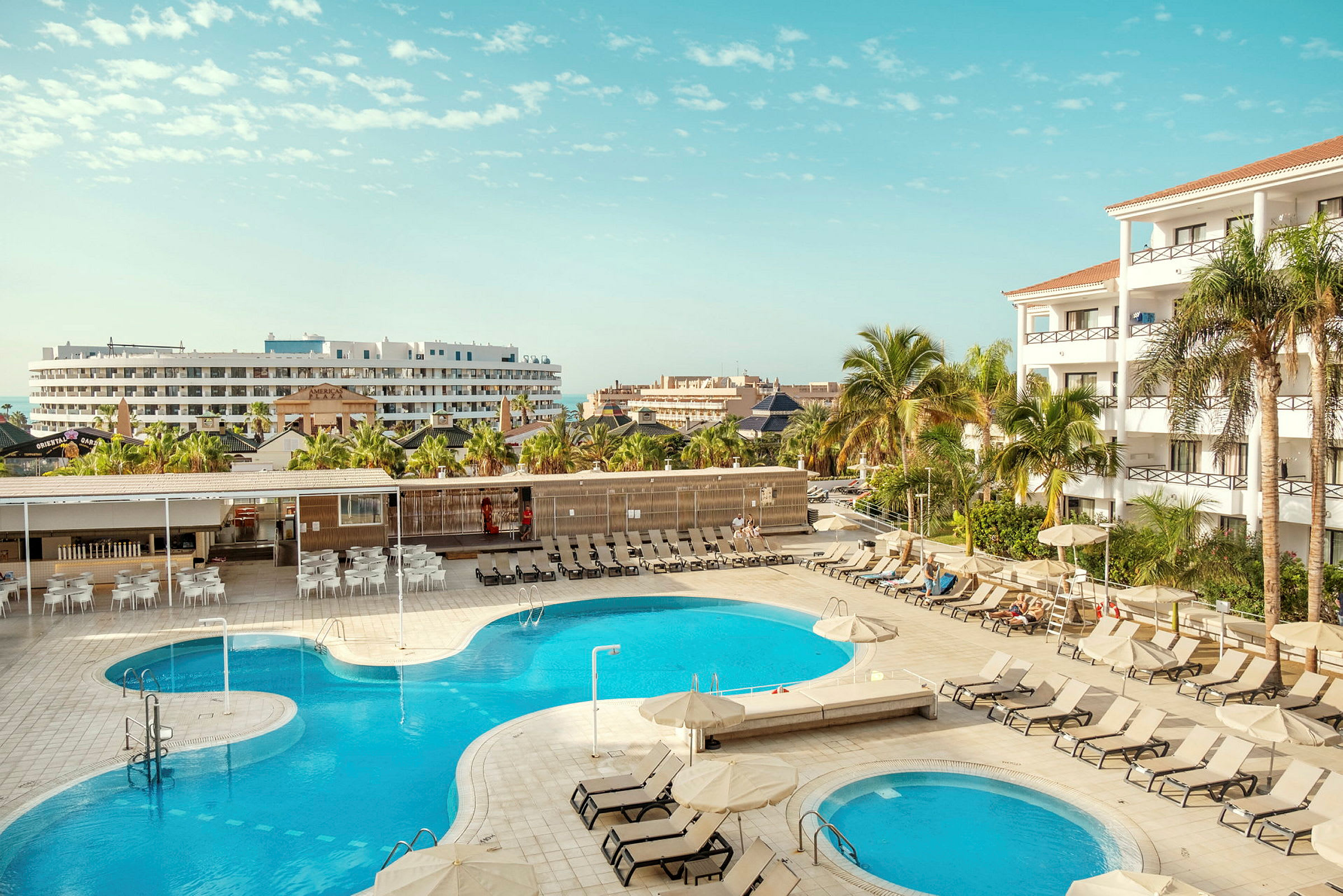 En hotelpool med solstole omkring og bygninger i baggrunden. (AI-genereret tekst)