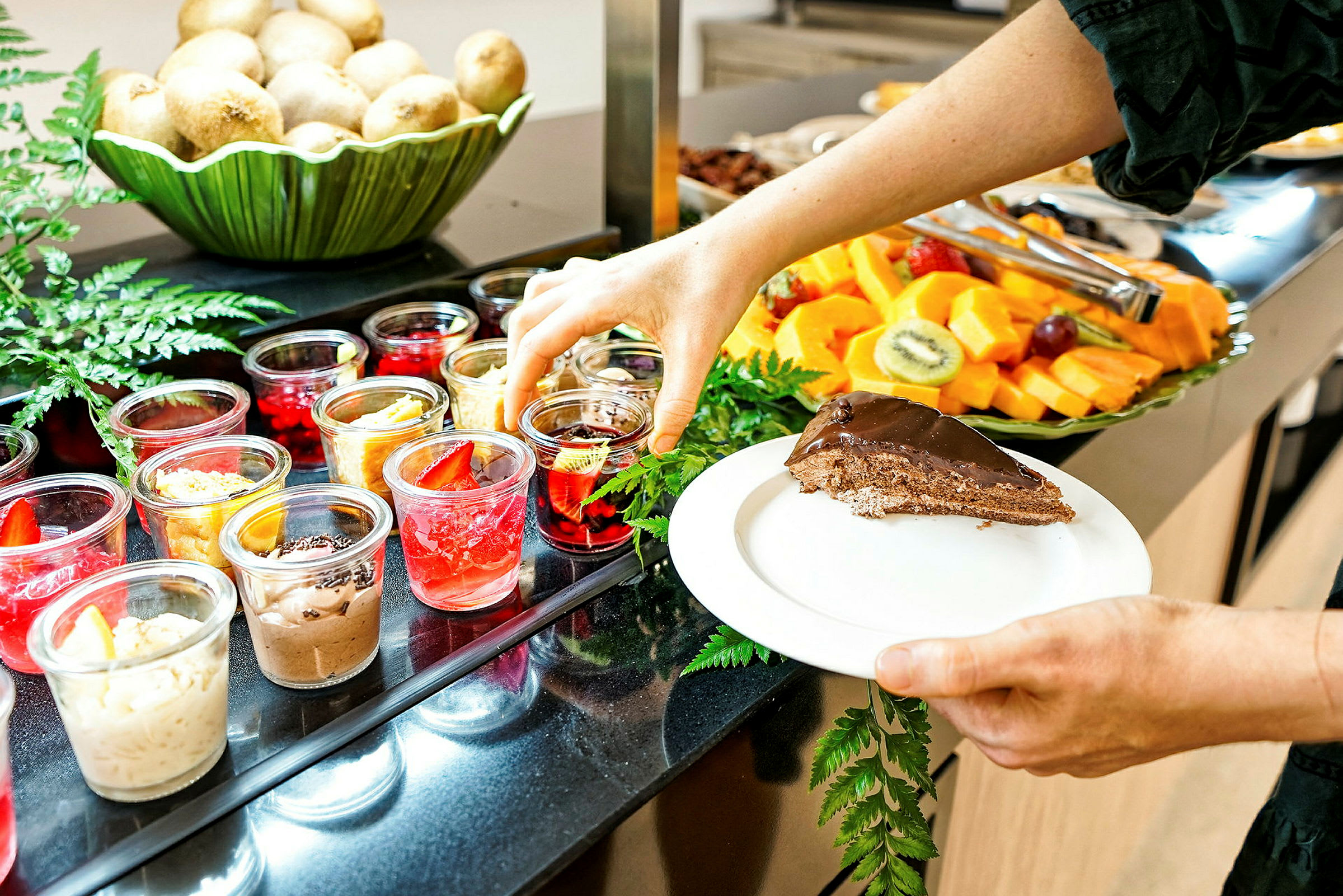 En person tager en dessert fra en buffet med et udvalg af små desserter i glas, frugter og et stykke chokoladekage på en tallerken. (AI-genereret tekst)