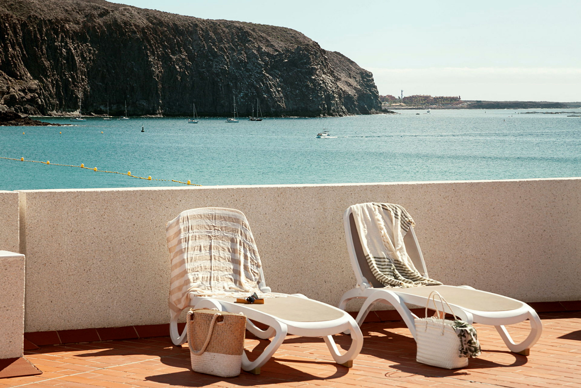 To liggestole med håndklæder og kurve placeret på en terrasse med udsigt over havet og en klippefyldt kyst. (AI-genereret tekst)