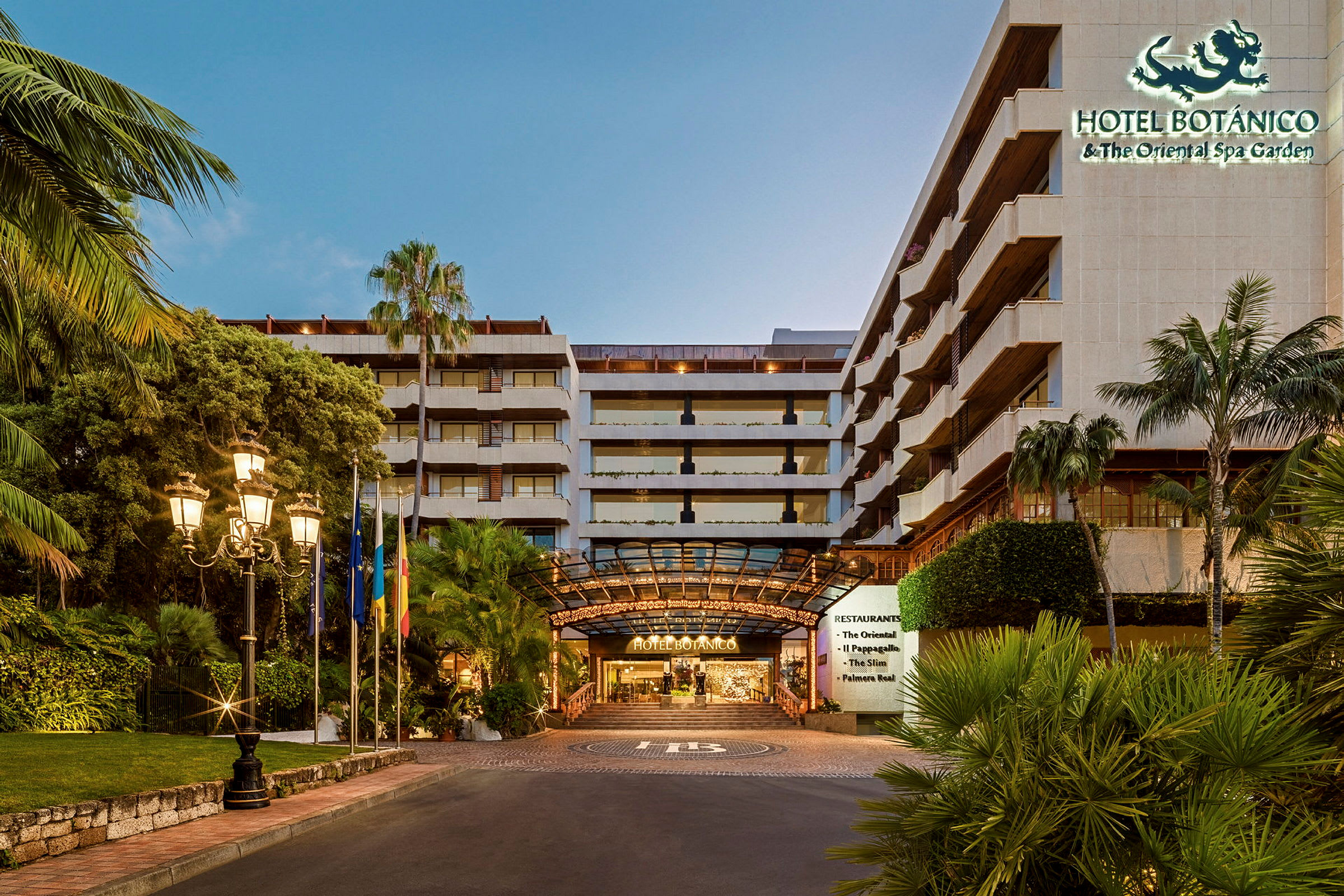 Indgangen til Hotel Botanico, omgivet af tropiske planter og oplyst af gadelamper. (AI-genereret tekst)