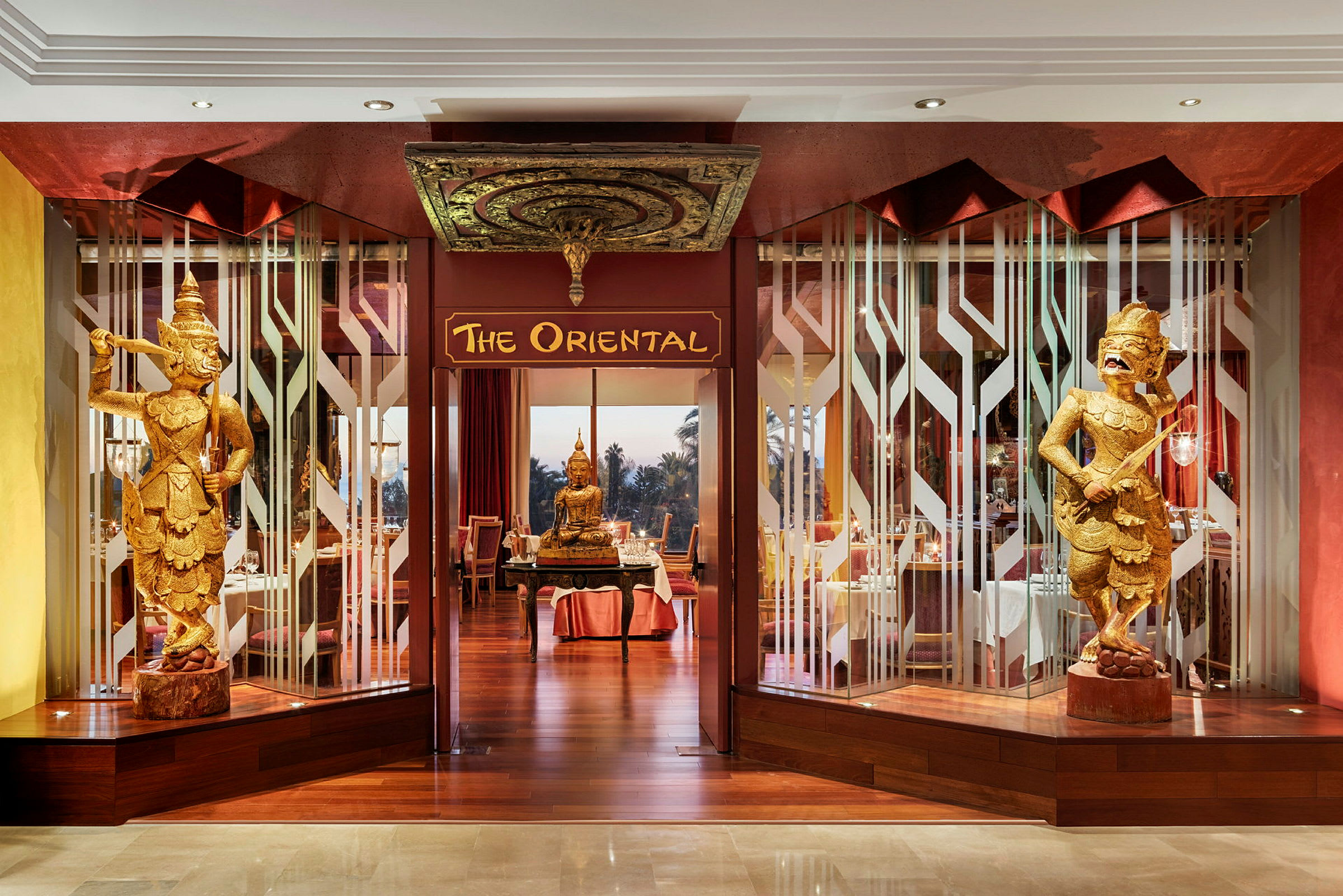 Indgangen til restauranten 'The Oriental' med dekorative gyldne figurer og en Buddha-statue. (AI-genereret tekst)