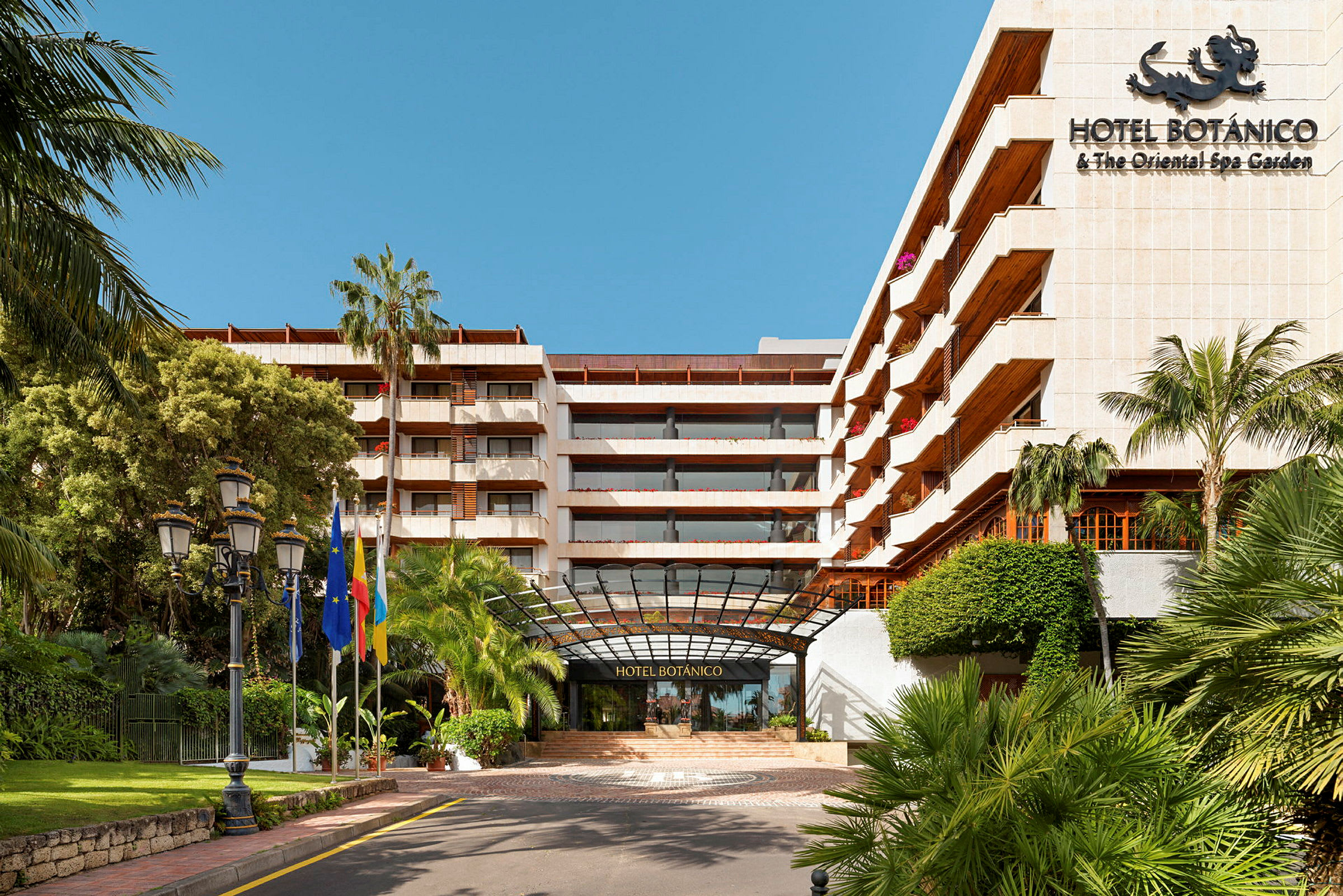 Hotellets facade med palmer og flag ved indgangen. (AI-genereret tekst)