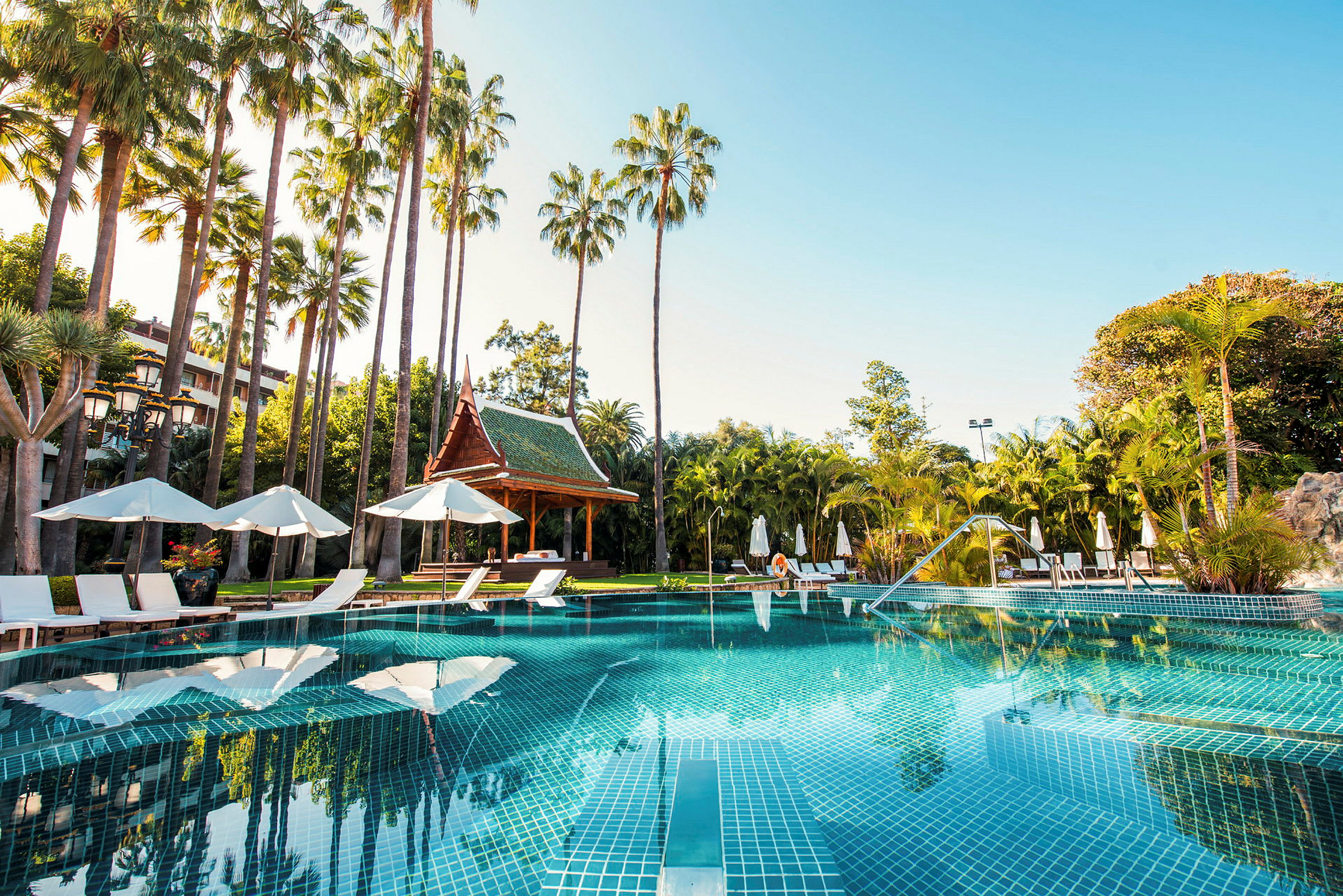 En swimmingpool omgivet af liggestole, parasoller og palmer med en thailandsk pavillon i baggrunden. (AI-genereret tekst)