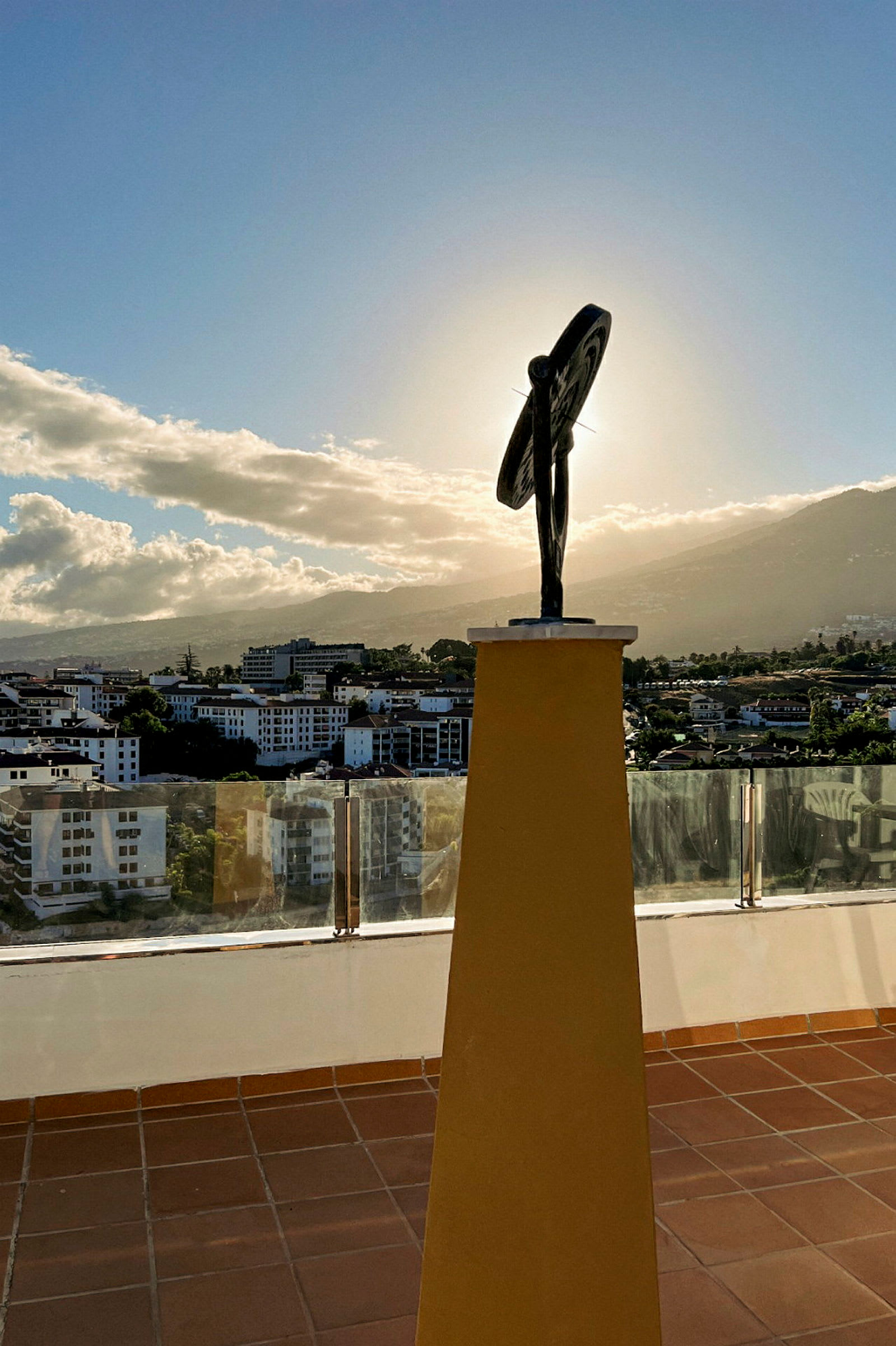 En skulptur på en gul piedestal med en by og bjerge i baggrunden. (AI-genereret tekst)