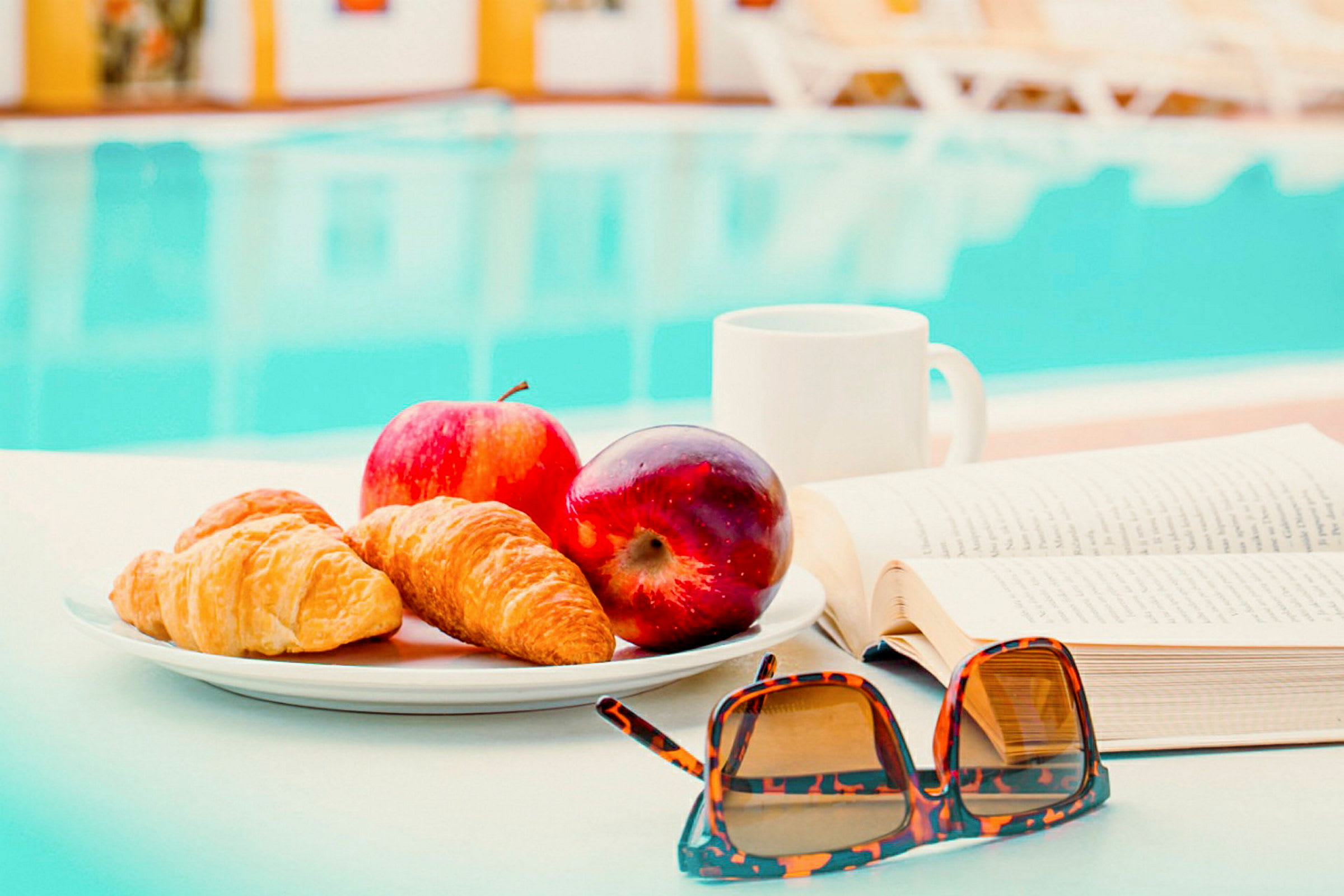 En tallerken med croissanter og æbler står på et bord ved siden af en bog og et par solbriller. I baggrunden er en pool synlig. (AI-genereret tekst)
