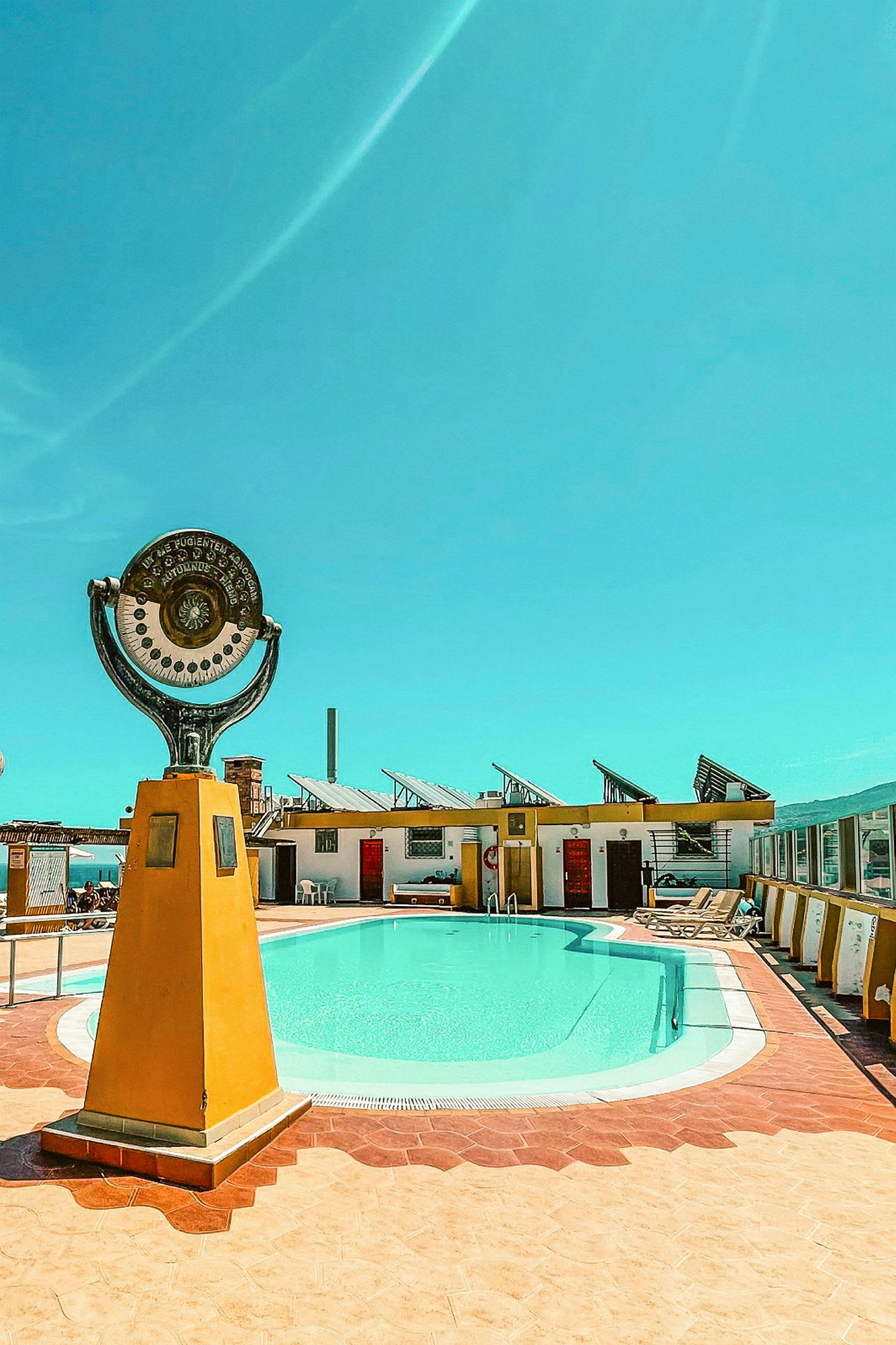 En udendørs swimmingpool med en dekorativ statue foran og bygninger omkring under en klar blå himmel. (AI-genereret tekst)
