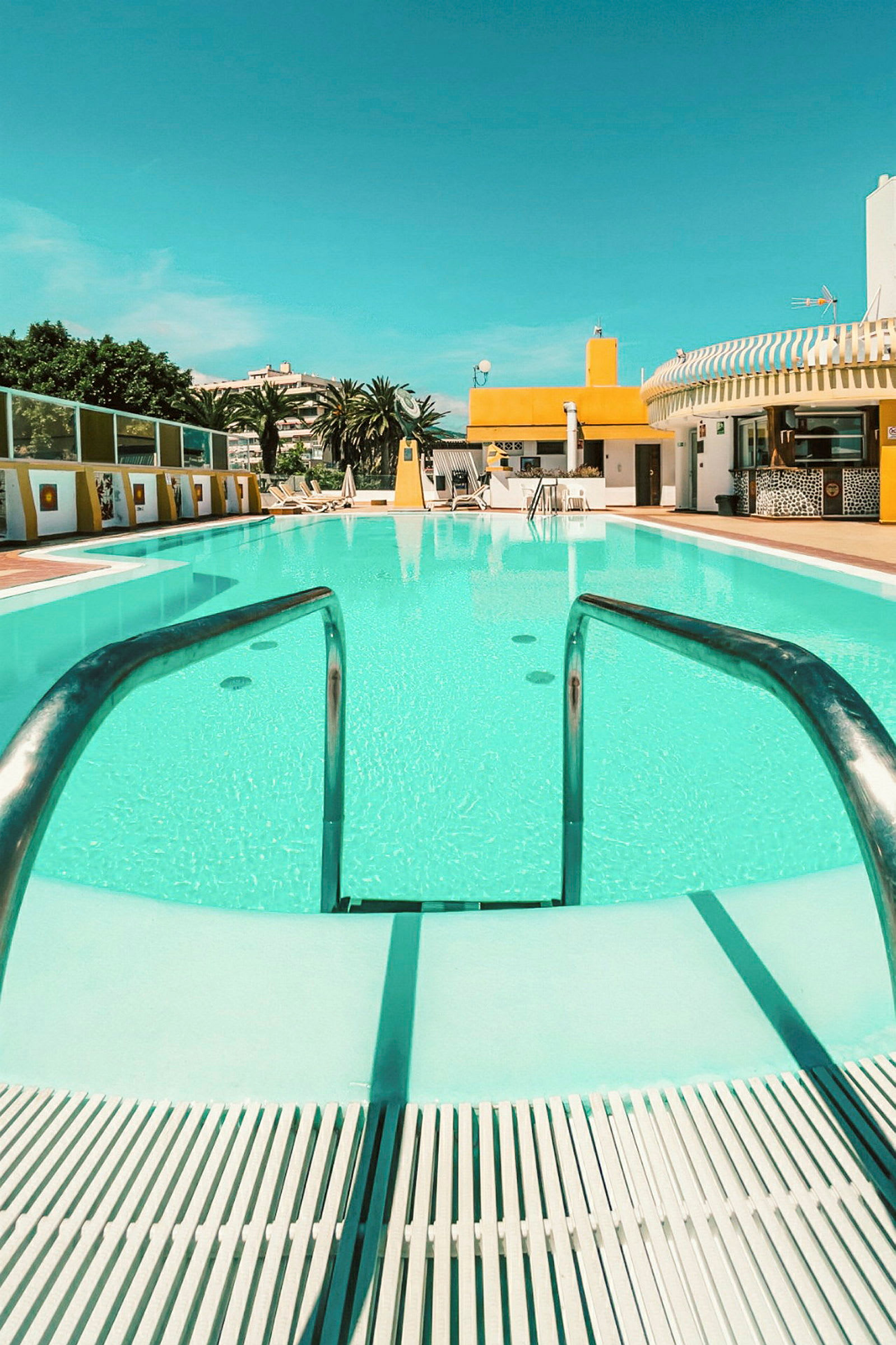 En pool med klart vand, omgivet af liggestole og en bygning med gul facade. (AI-genereret tekst)