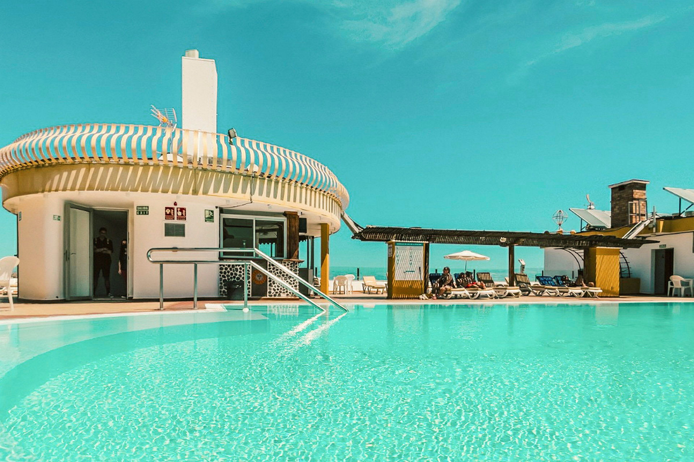 En udendørs swimmingpool ved en bygning med liggestole og en klar blå himmel. (AI-genereret tekst)