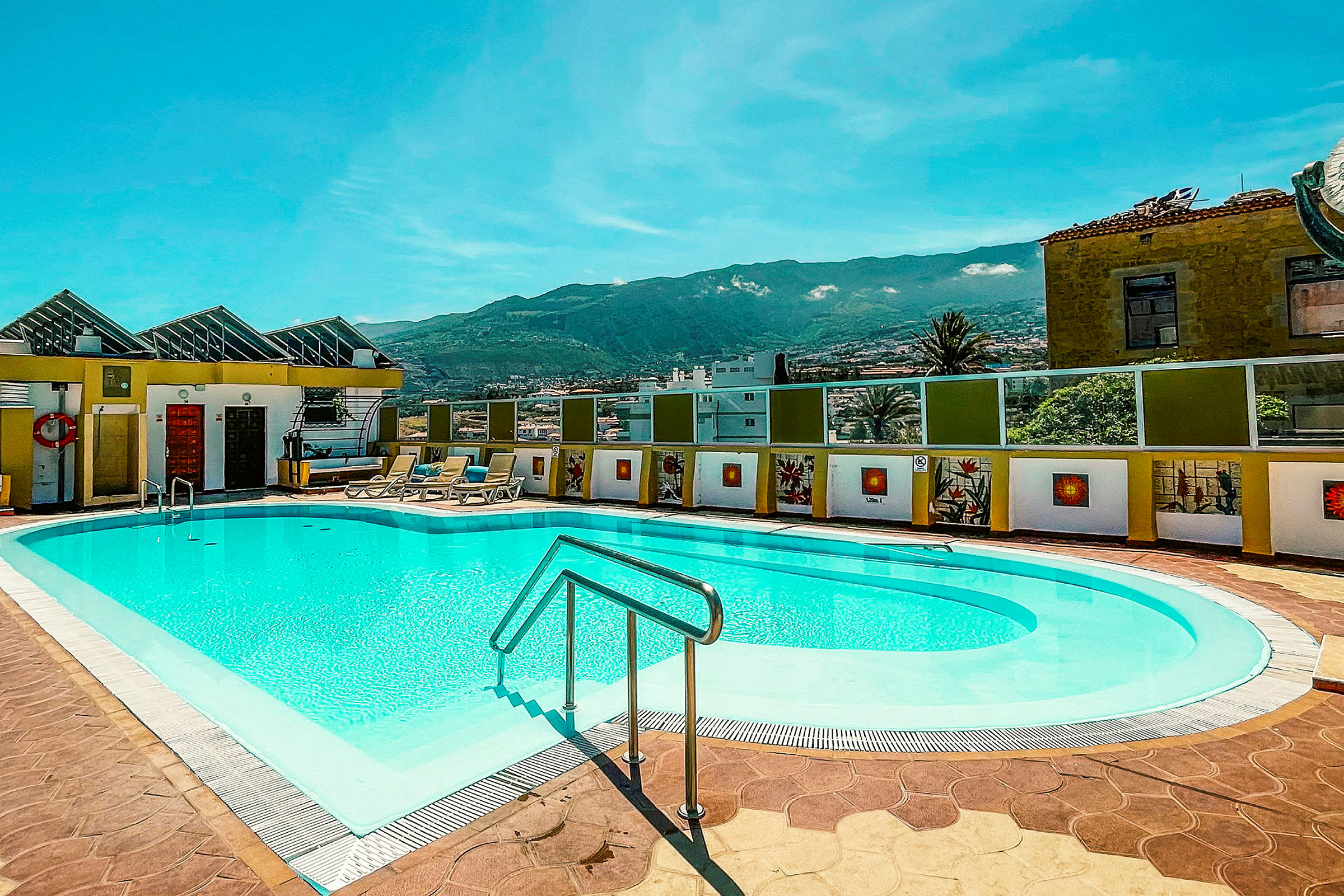Udendørs swimmingpool med liggestole i en solrig atmosfære, omgivet af bygninger og med bjerge i baggrunden. (AI-genereret tekst)