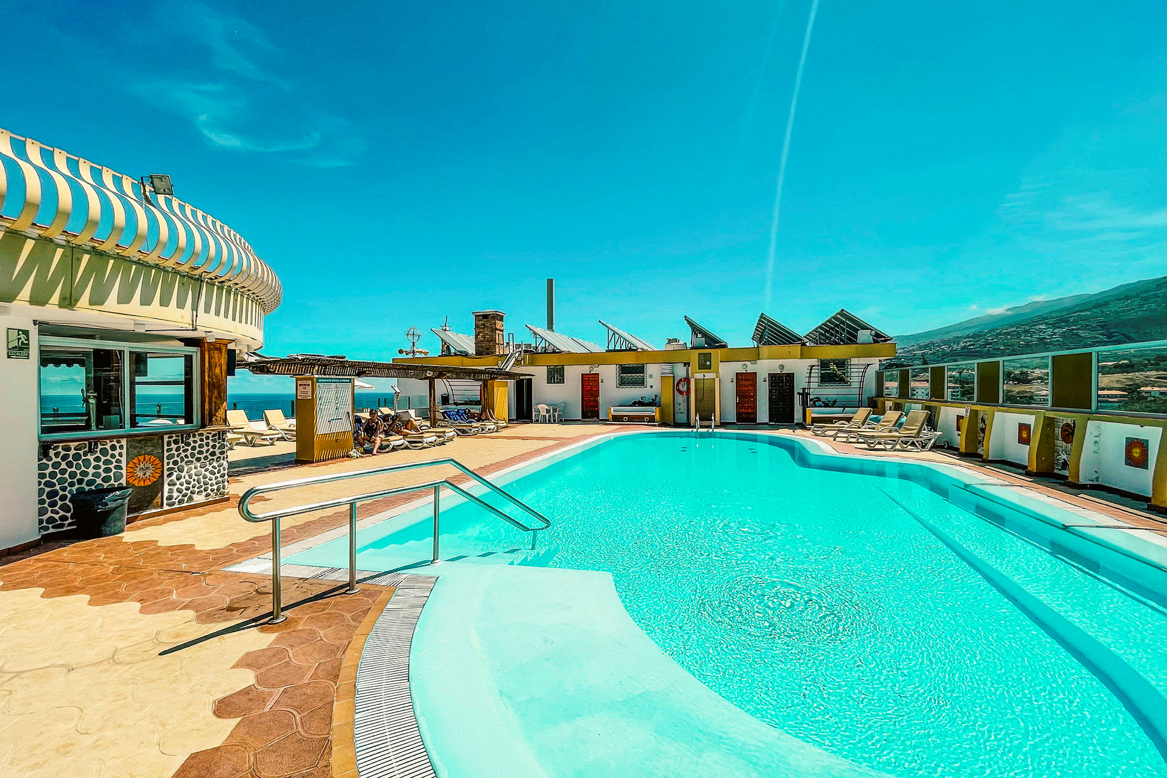 En udendørs swimmingpool med klart blåt vand, omgivet af liggestole og bygninger under en klar blå himmel. (AI-genereret tekst)