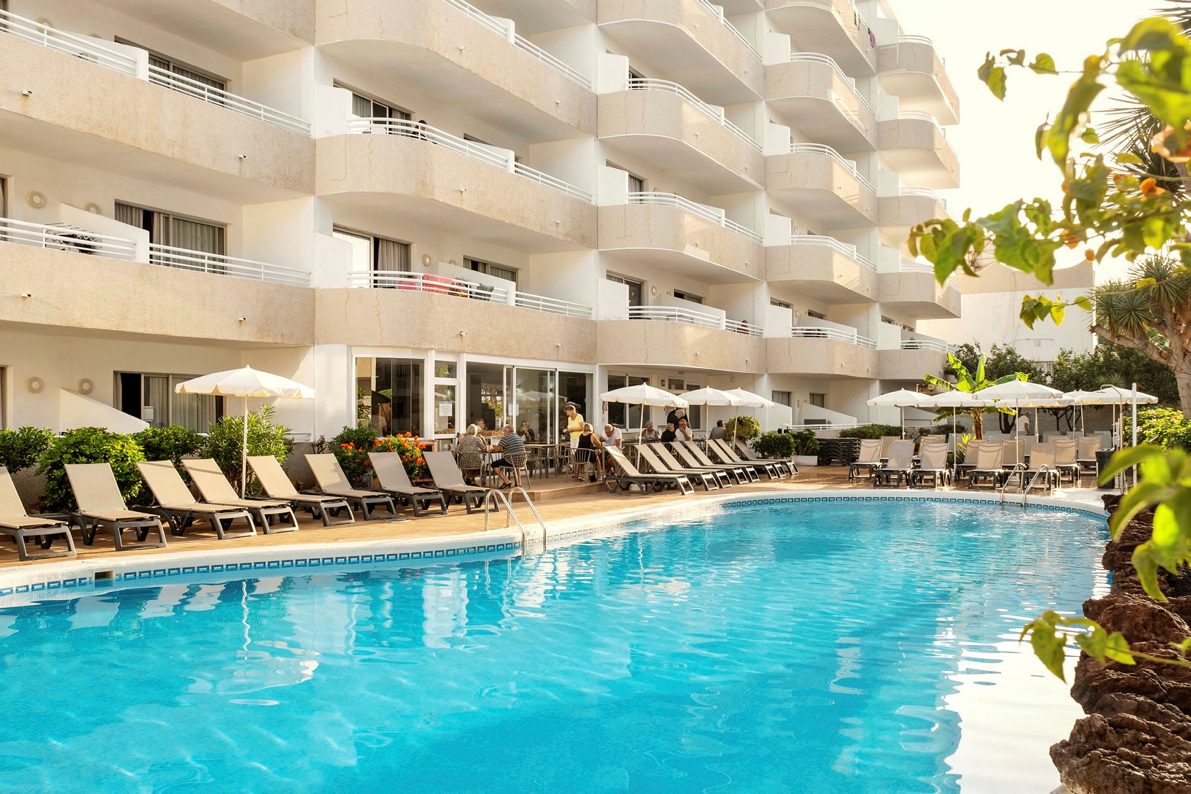 Billede af et hotel med altaner, pool og liggestole. (AI-genereret tekst)